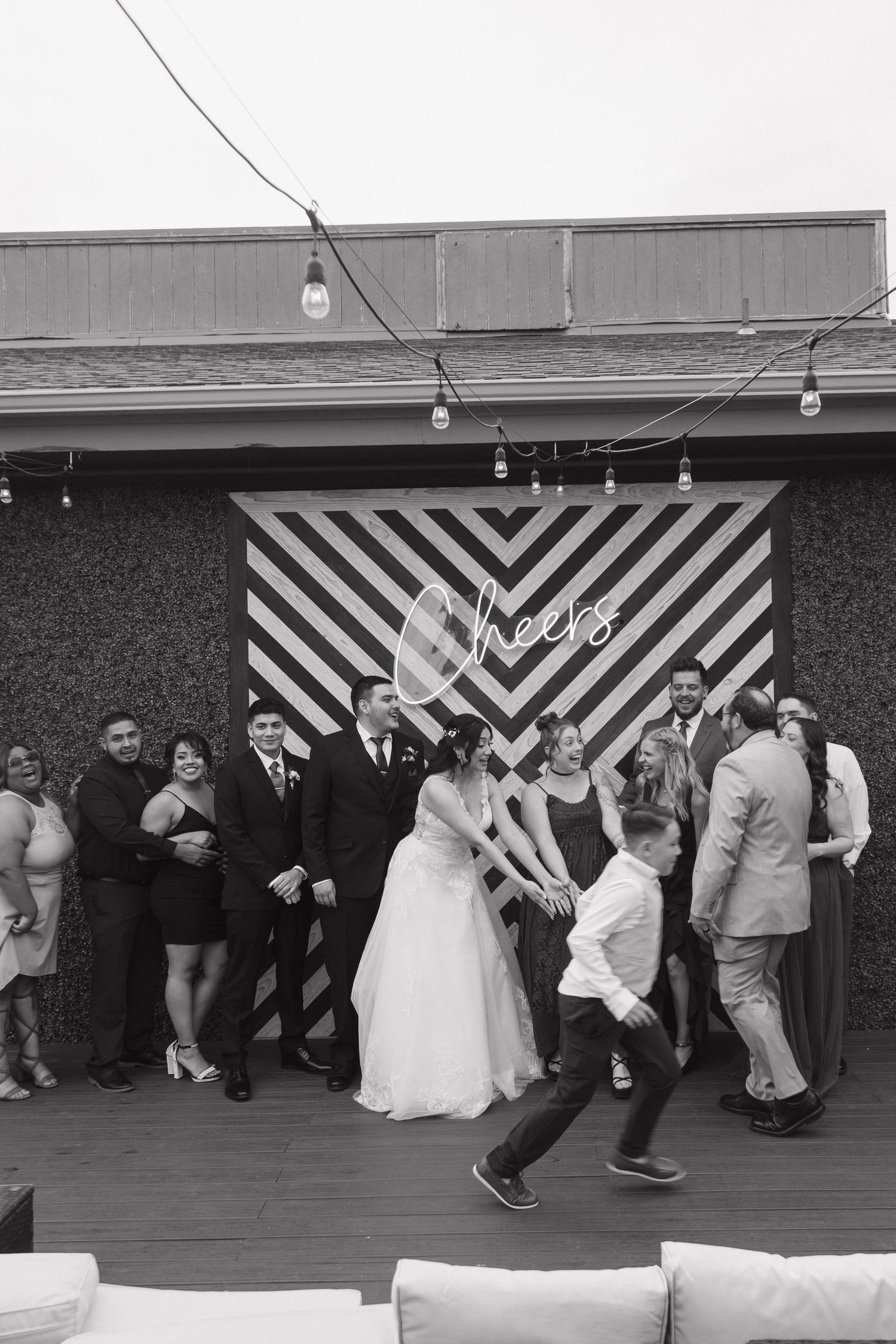 Black and white wedding photo of couple and guests celebrating in front of a Cheers backdrop.