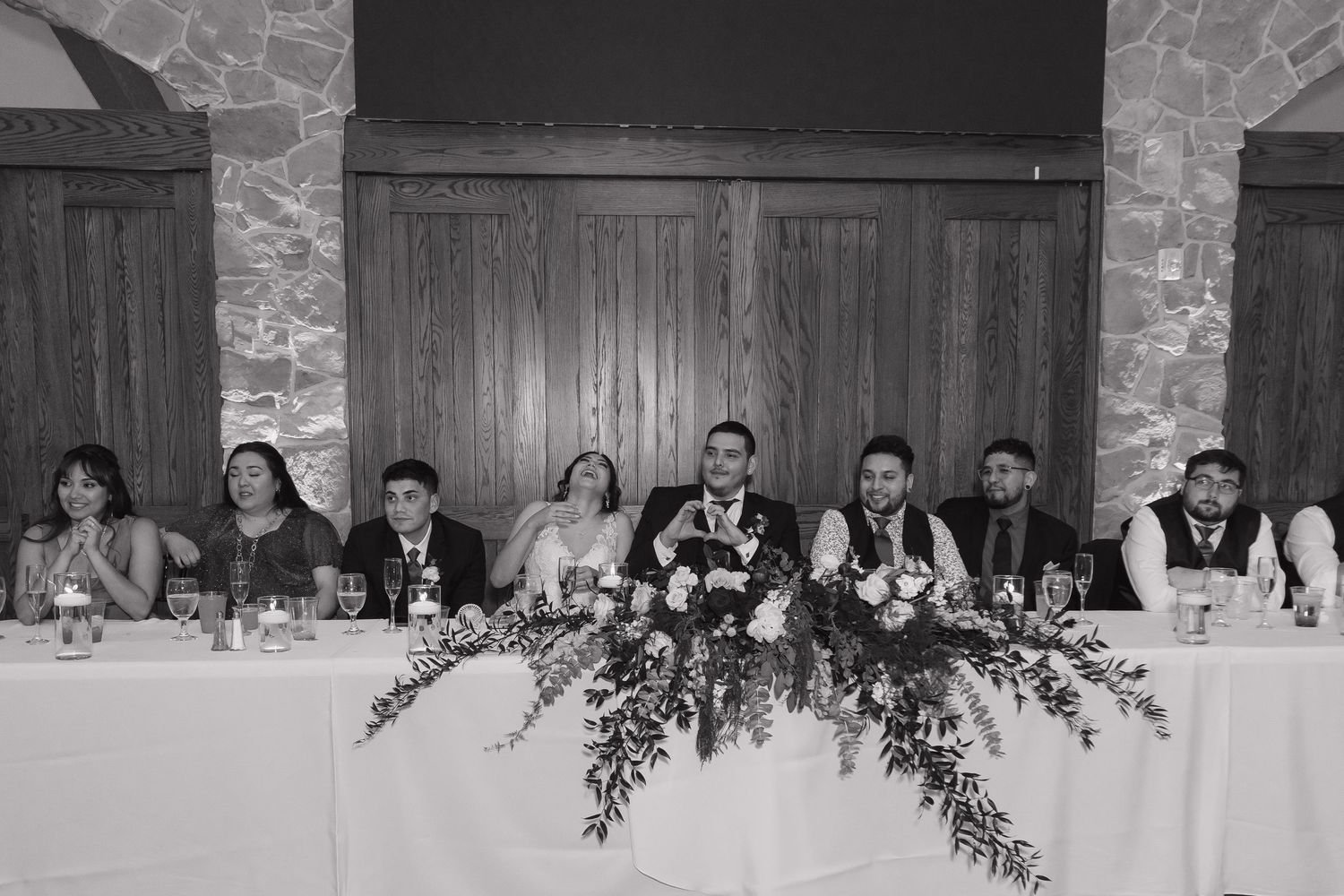 Black and white wide shot of wedding party seated at head table with lush floral centerpiece at reception.