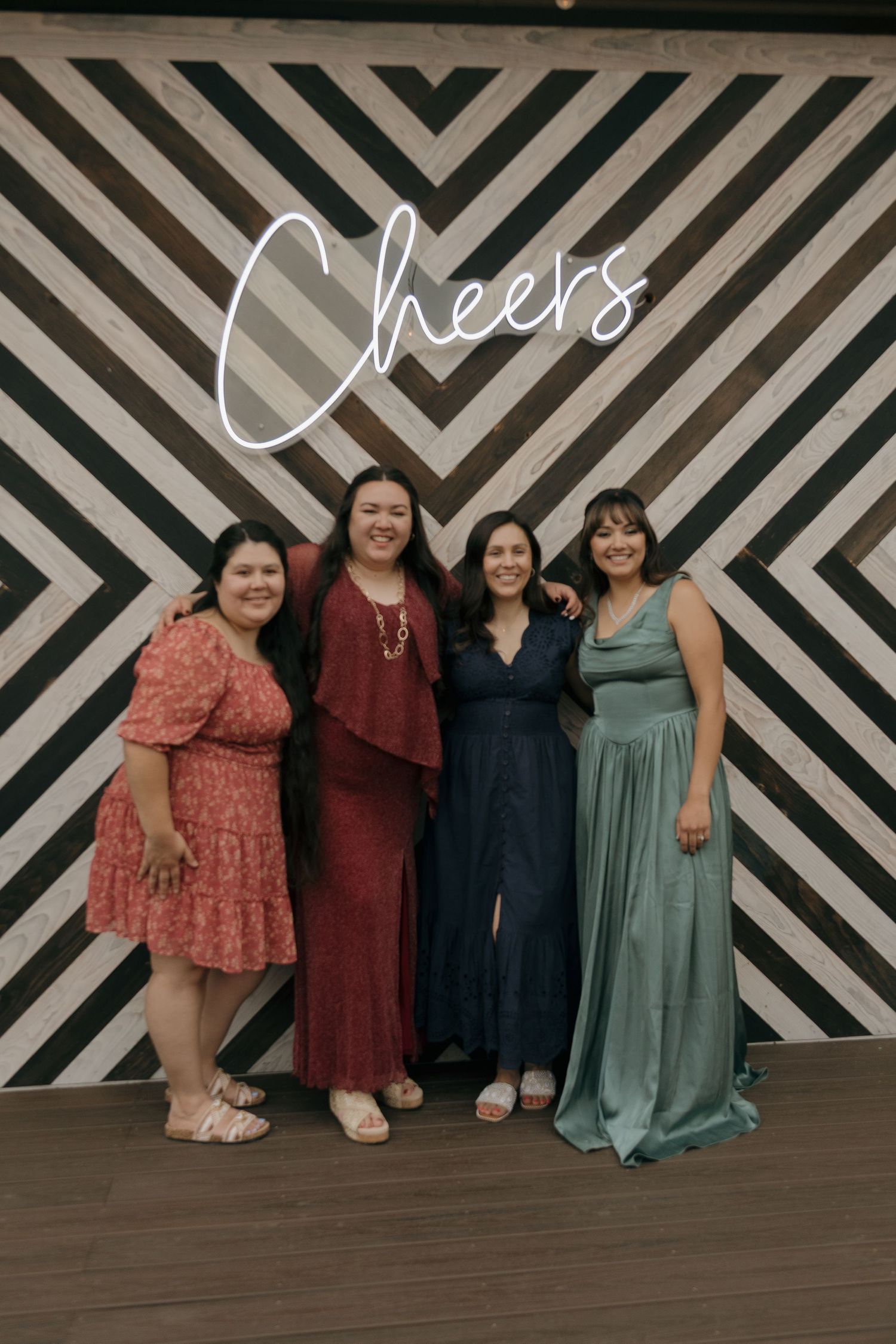Four women in elegant dresses pose in front of a Cheers neon sign at a wedding.