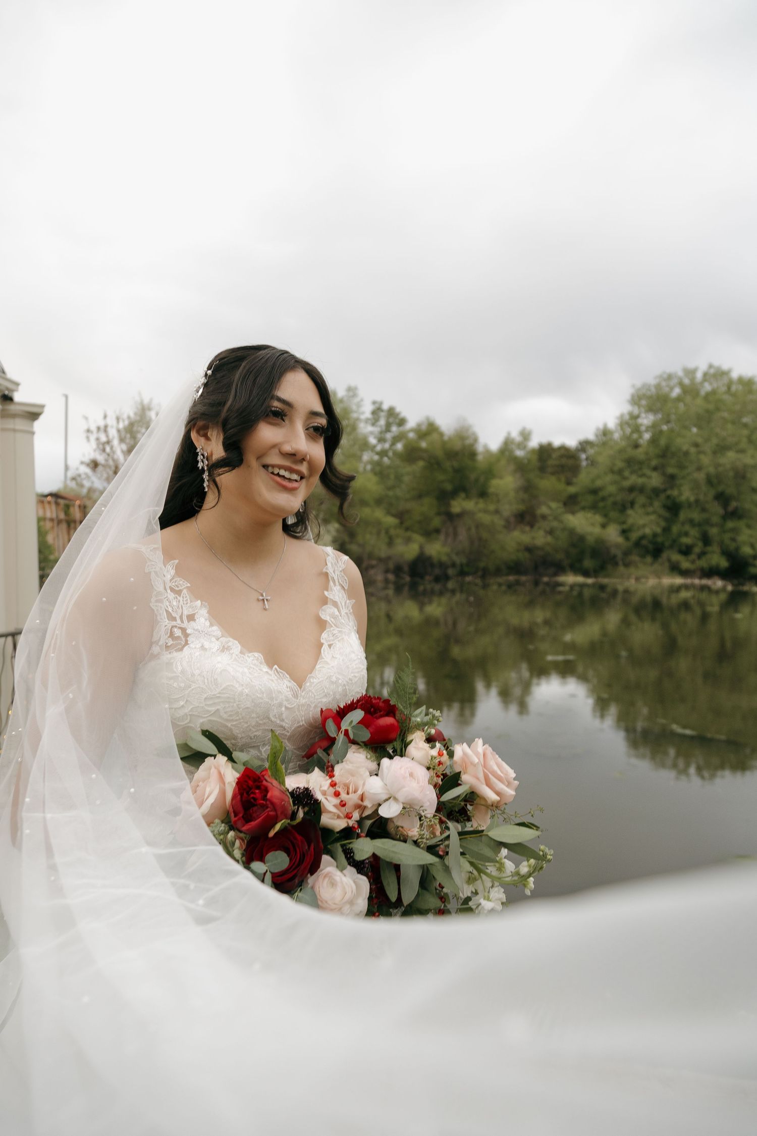 Smiling bride in lace wedding gown holding red and pink bouquet with veil flowing near a serene lake.