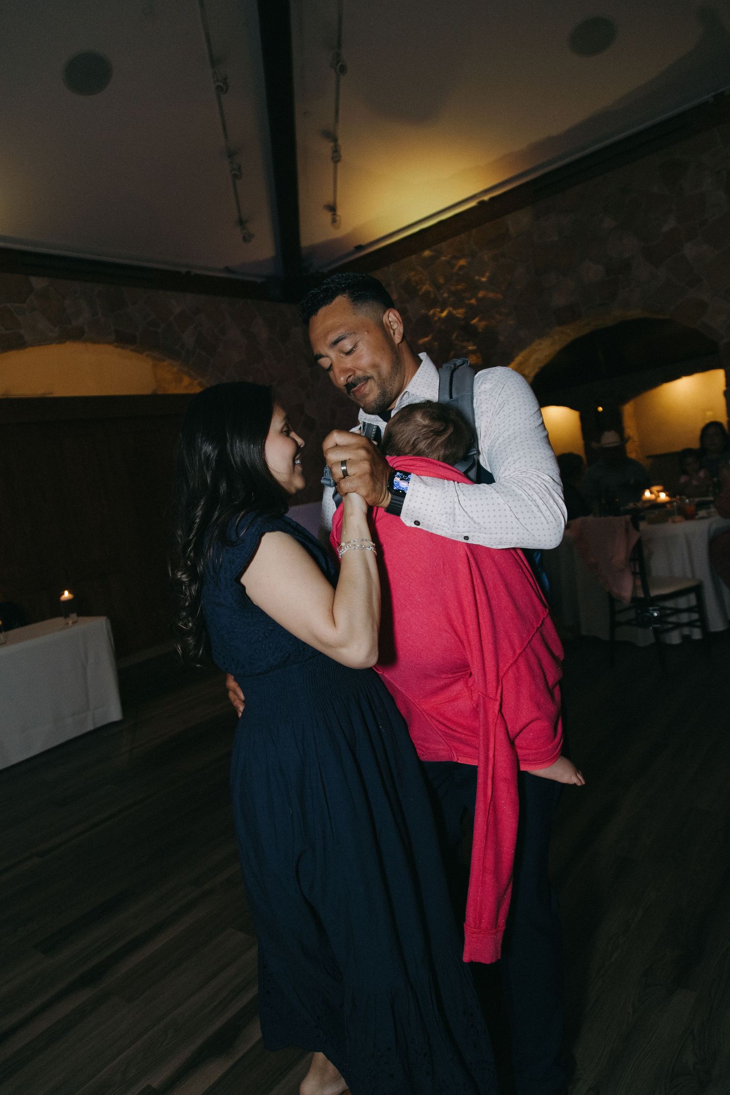 Happy couple shares a dance while groom carries their newborn baby in a red carrier at a wedding reception.