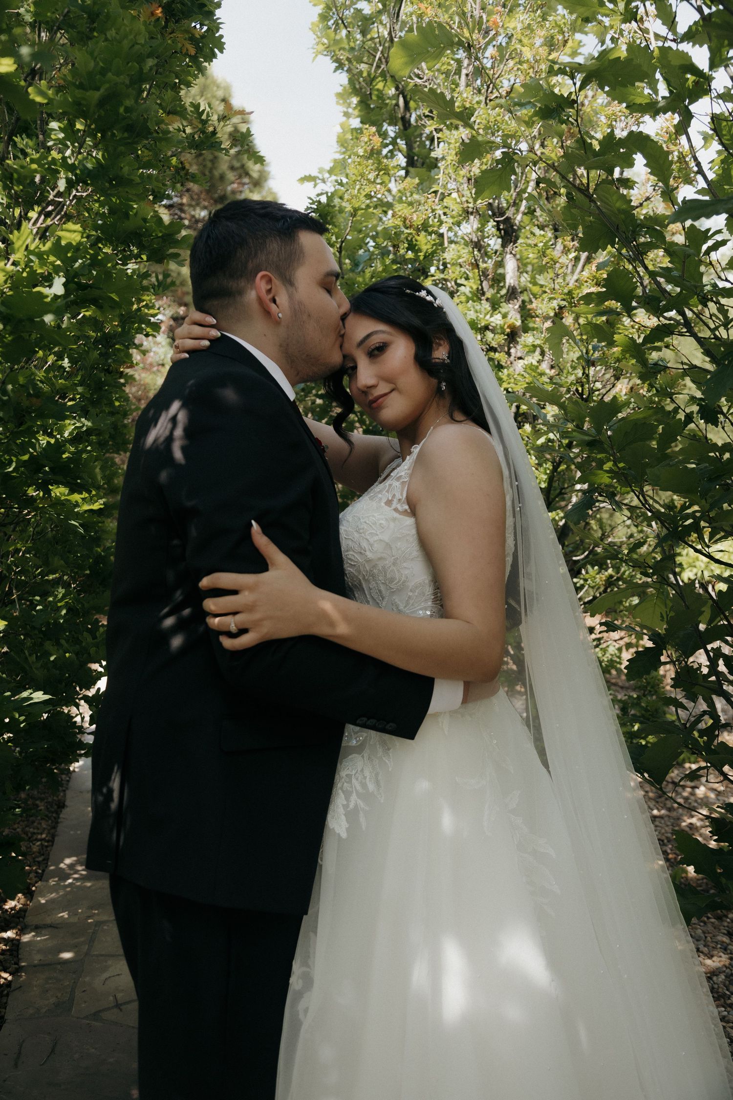 Groom kissing brides forehead as she smiles in lace wedding gown among green trees.