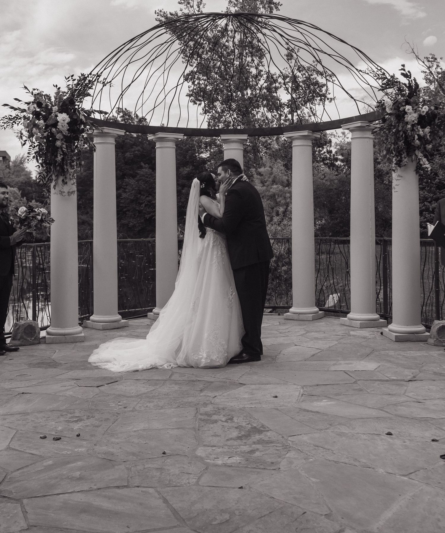 Black and white photo of bride and groom sharing first kiss under a floral gazebo at their wedding.
