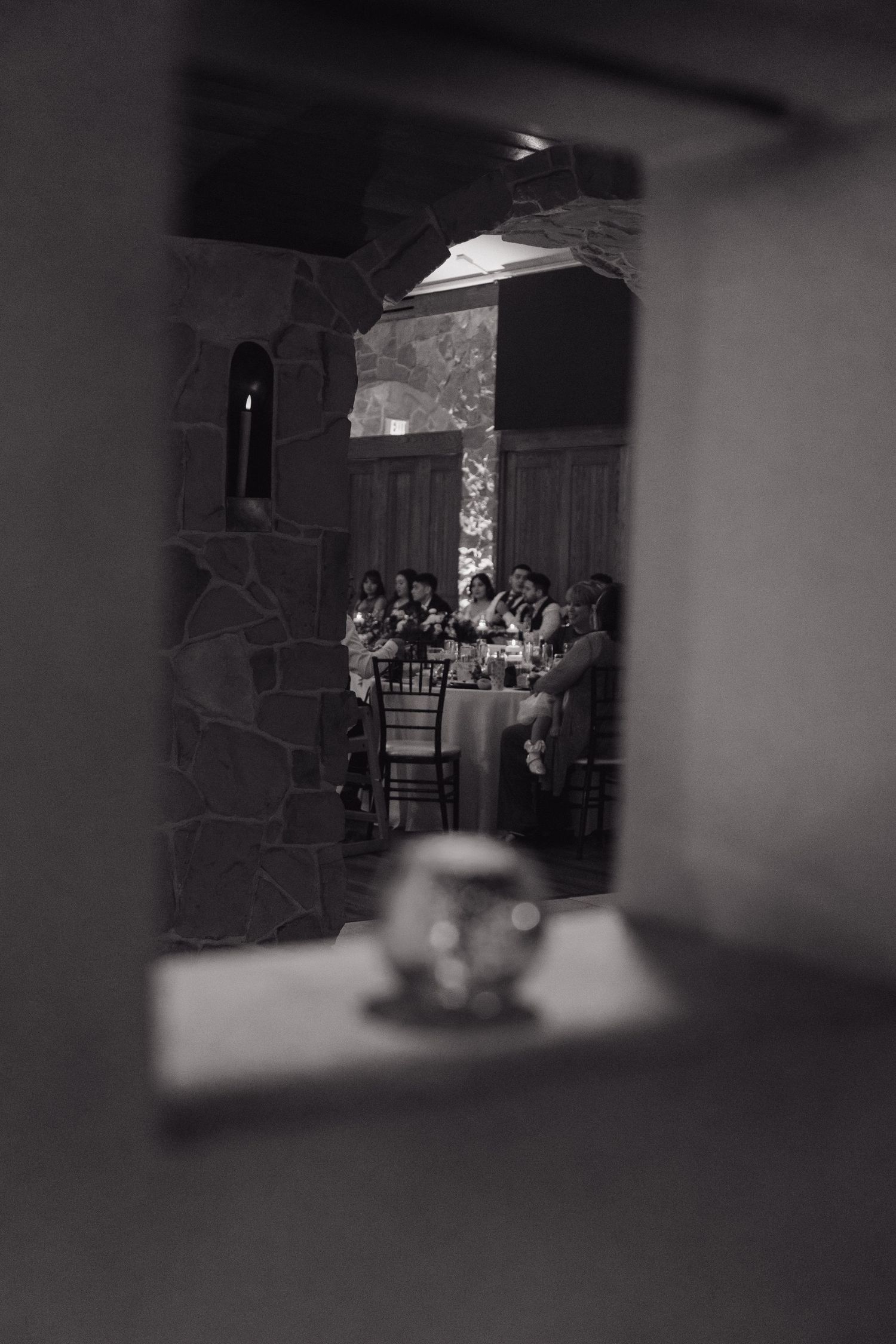 Black and white photo of wedding guests seated at tables viewed through a stone archway doorway.