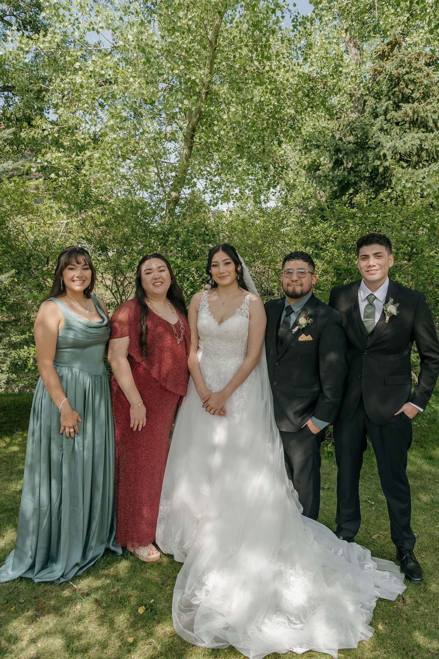 Bride in white lace gown poses with family members in formal attire under lush green trees outdoors.