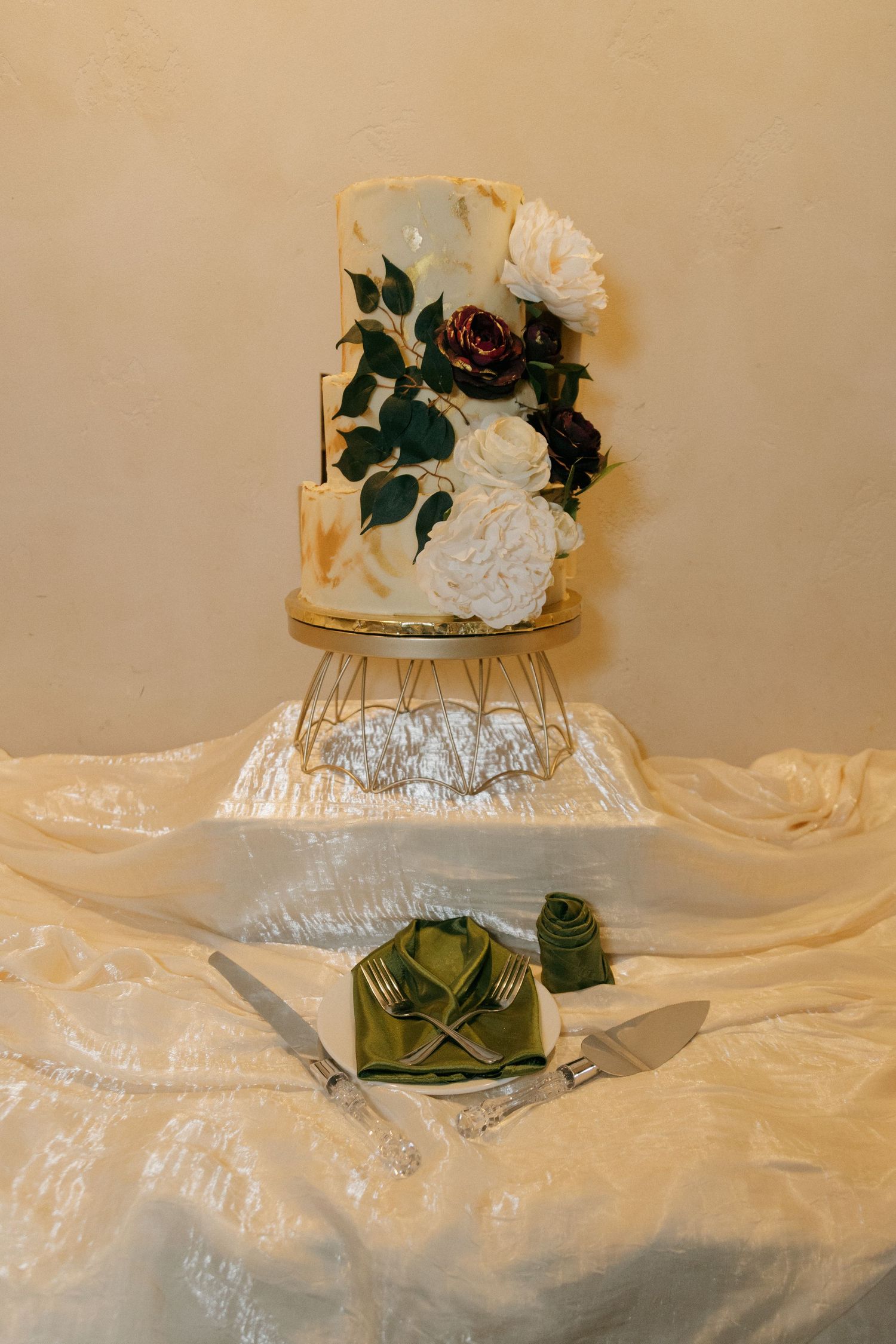 Three-tier wedding cake with white and burgundy floral decorations on a gold geometric stand.