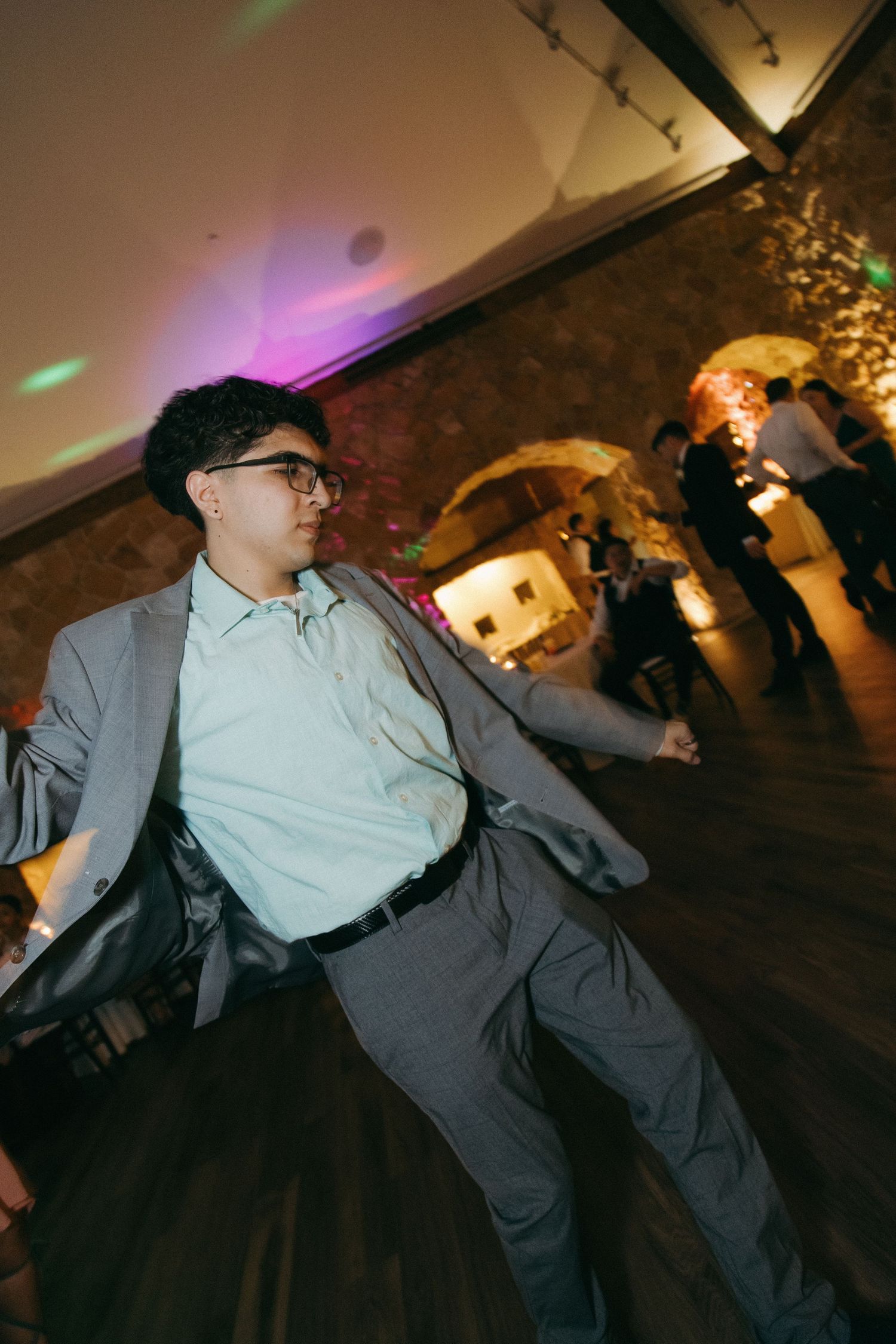 A young wedding guest in a gray suit and glasses dances at a warmly lit wedding reception venue.