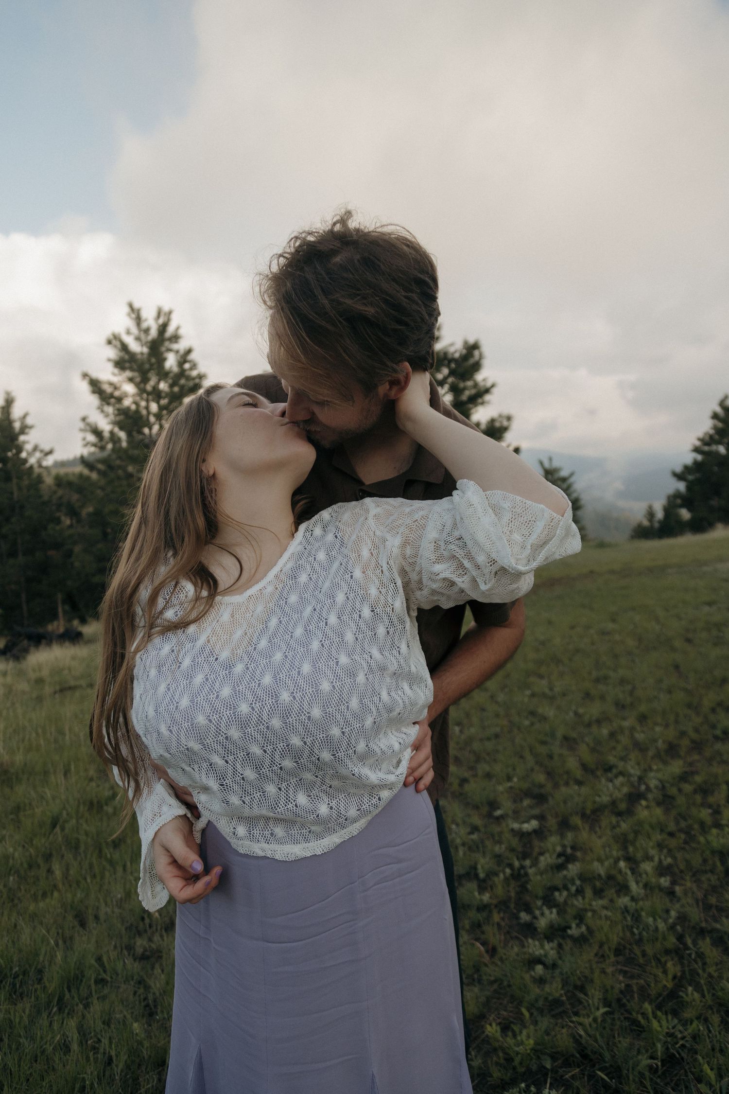 Young couple kissing passionately in a green meadow, woman in white lace top and lavender skirt.