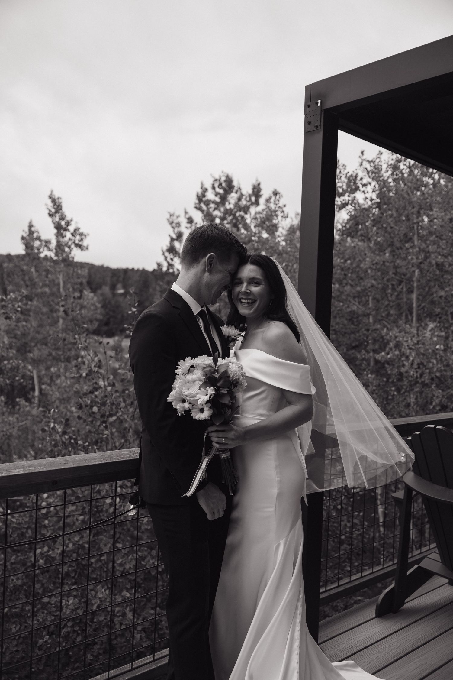 Black and white photo of bride and groom laughing together on wooden deck with forest backdrop.