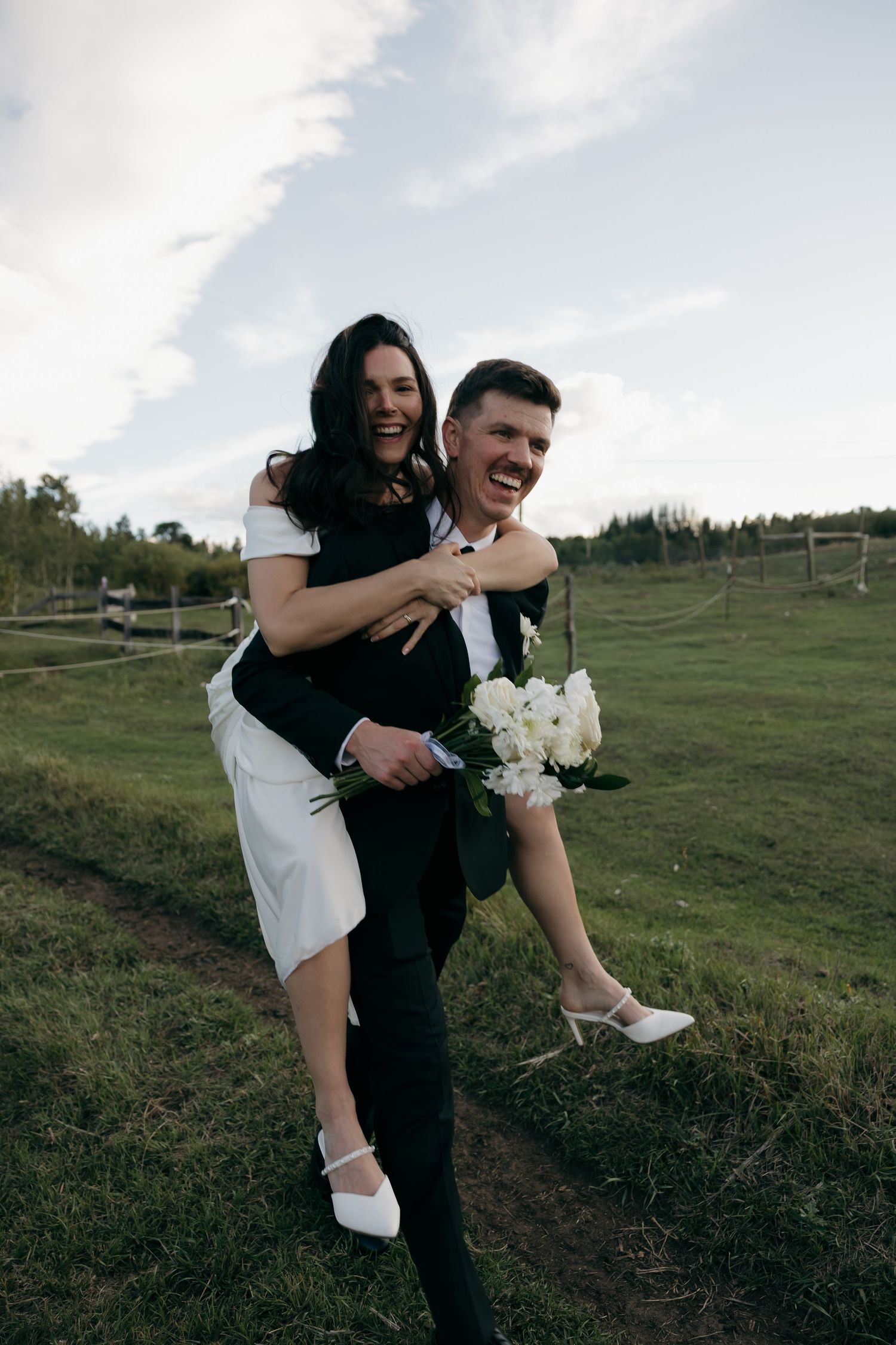 Laughing groom gives joyful bride a piggyback ride across green countryside field at wedding.