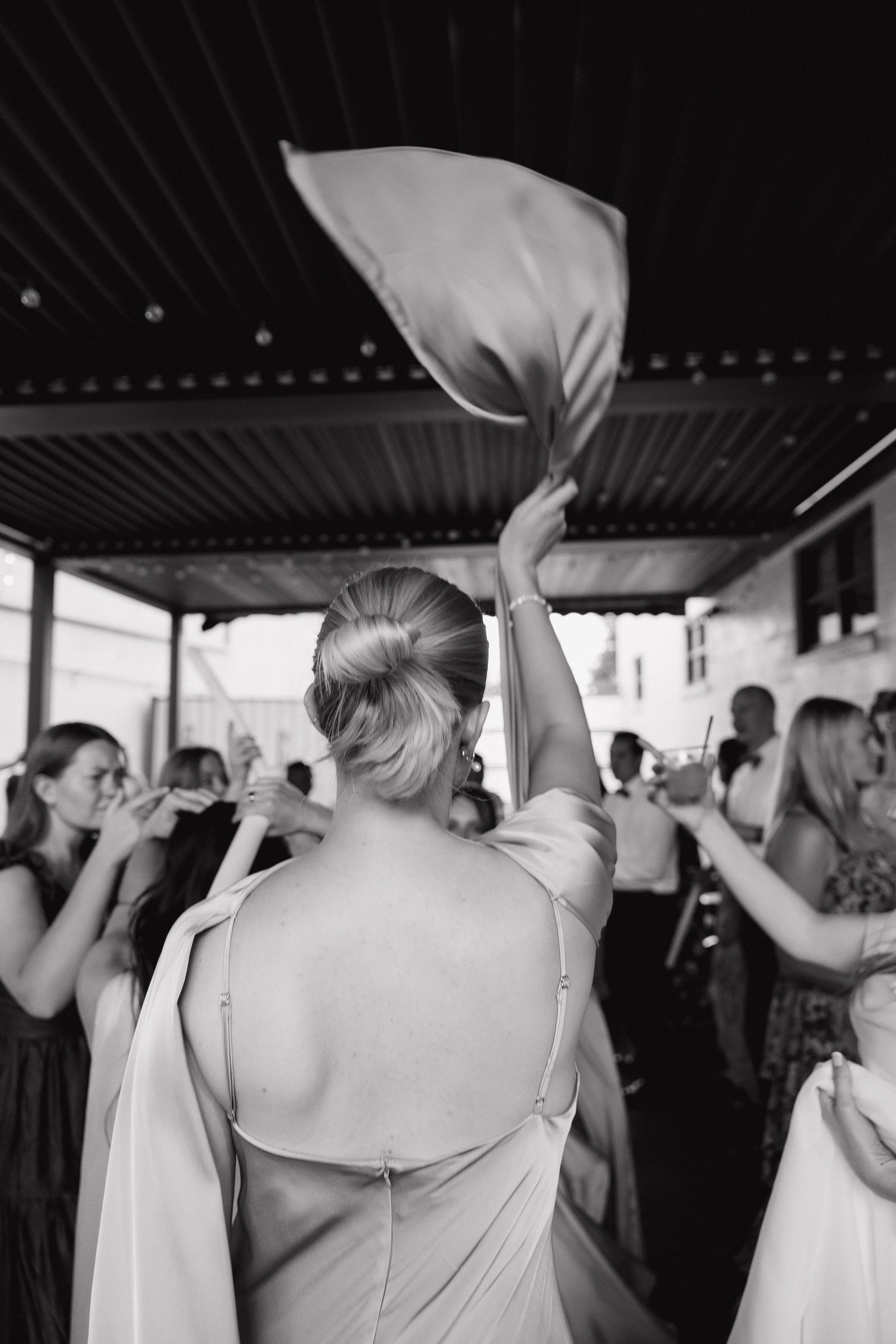 Bridesmaid in backless satin gown waves fabric in air during lively outdoor wedding reception celebration in black and white.
