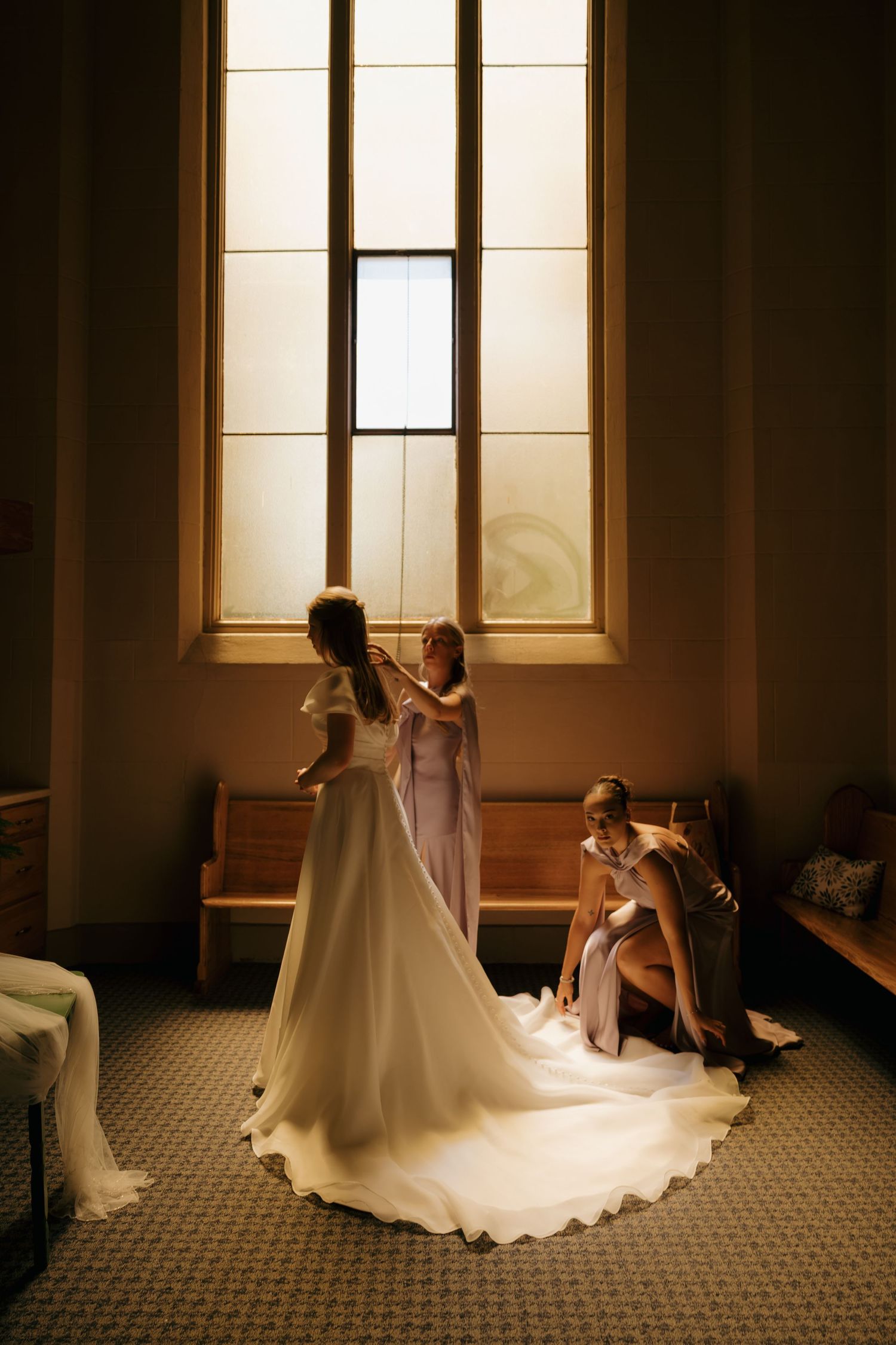 Bride in white gown with long train stands by stained glass window as bridesmaids help her prepare in a church.