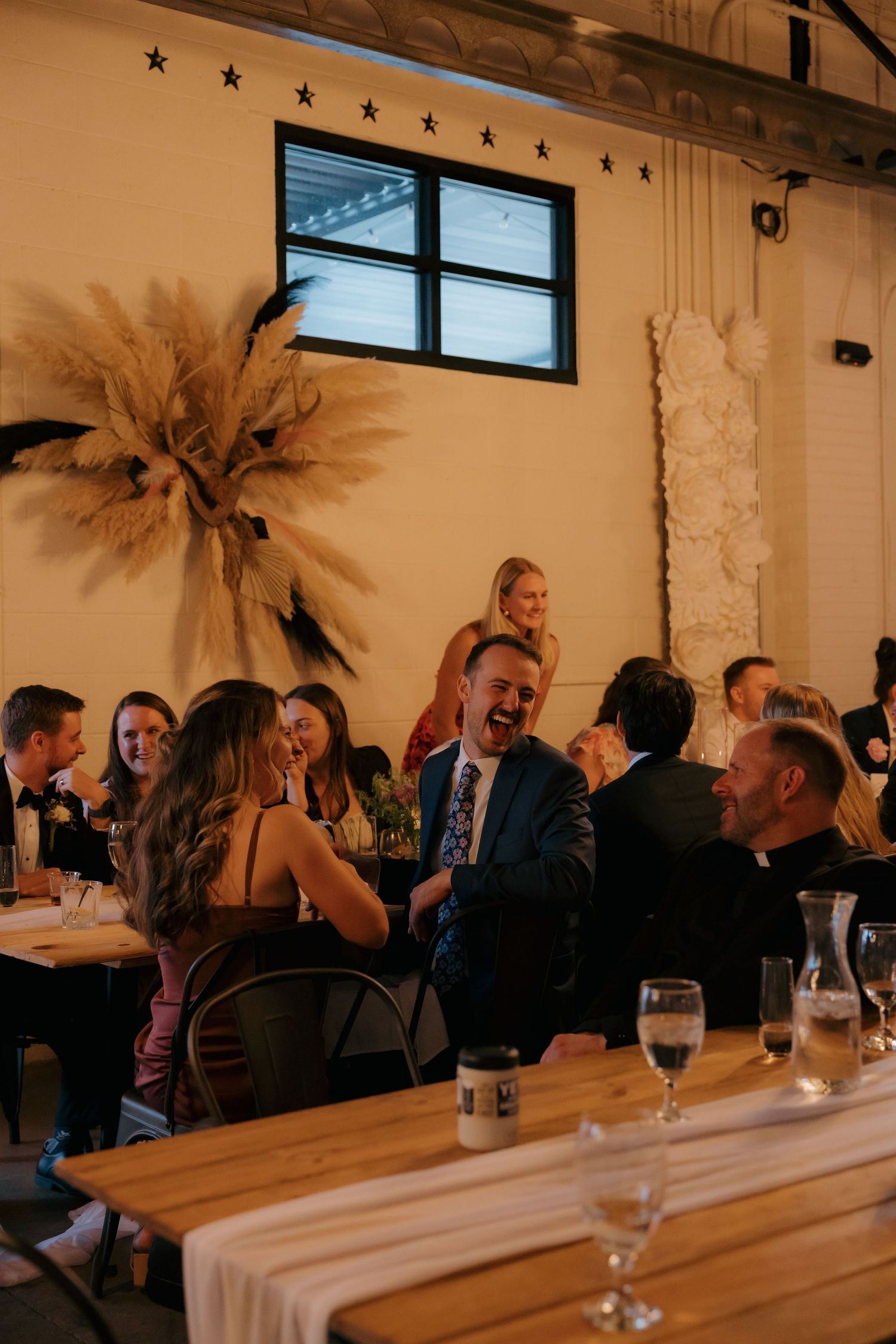 Wedding reception guests laughing and celebrating at wooden tables in industrial venue with pampas grass decor.