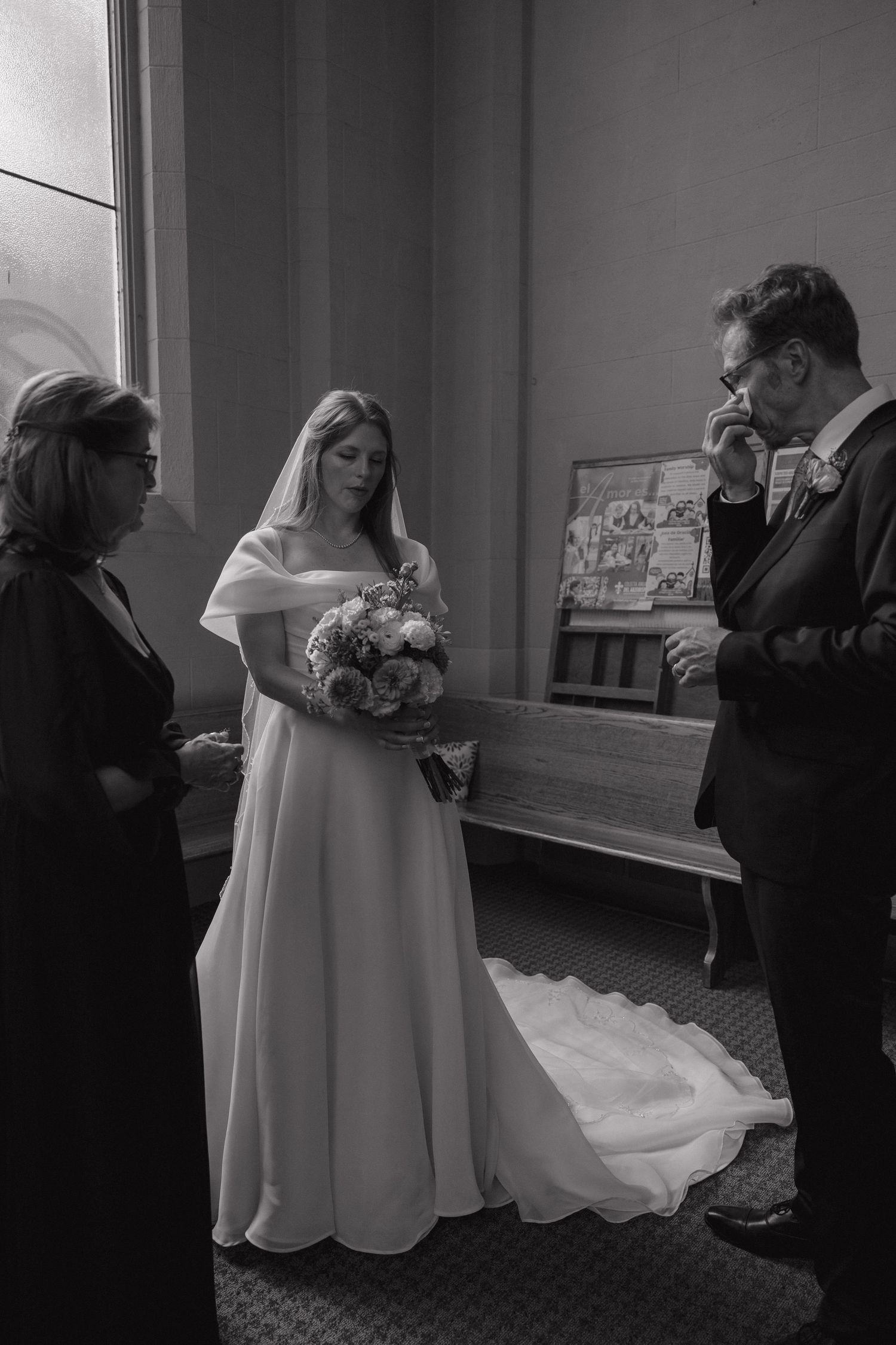 Black and white photo of bride in elegant gown holding bouquet as emotional groom wipes tears before wedding ceremony.