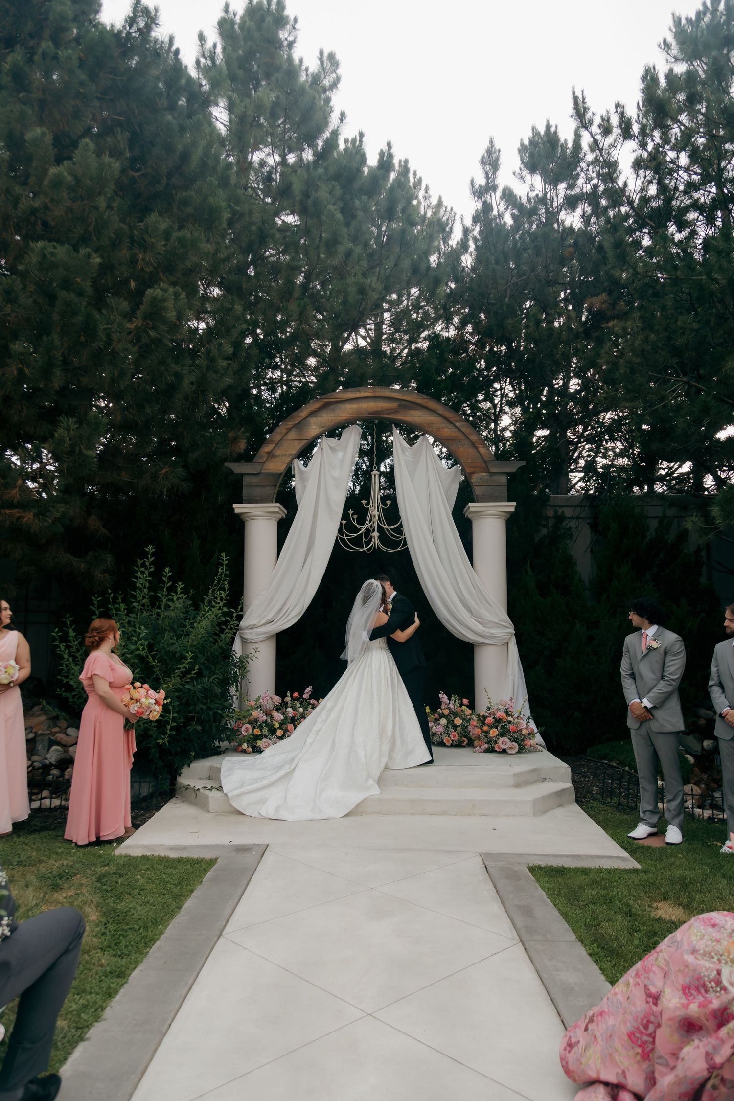 Bride and groom exchange vows under elegant arch with white draping and floral arrangements in outdoor garden wedding ceremony.