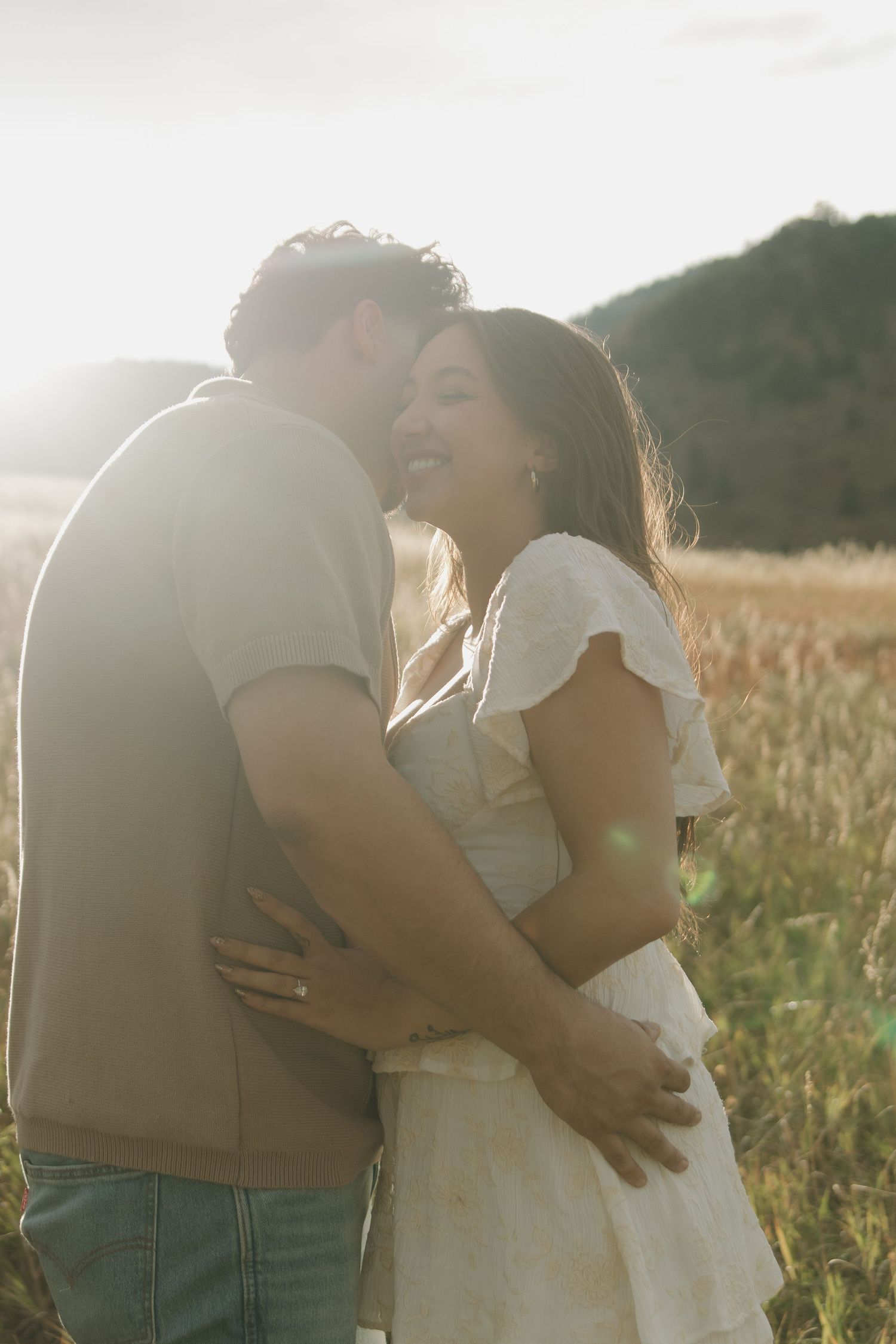 Couple embracing and laughing in sunlit field with warm golden backlight during engagement shoot.
