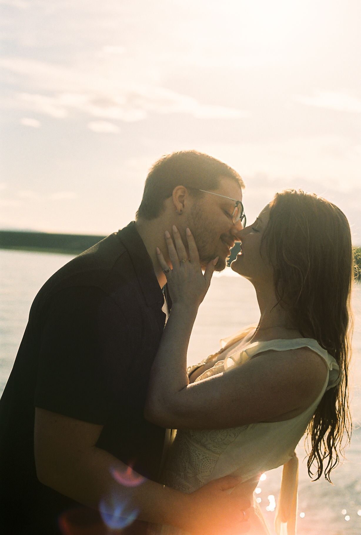 Couple sharing a passionate kiss at golden hour by a lake, sun flare creating romantic warm glow.