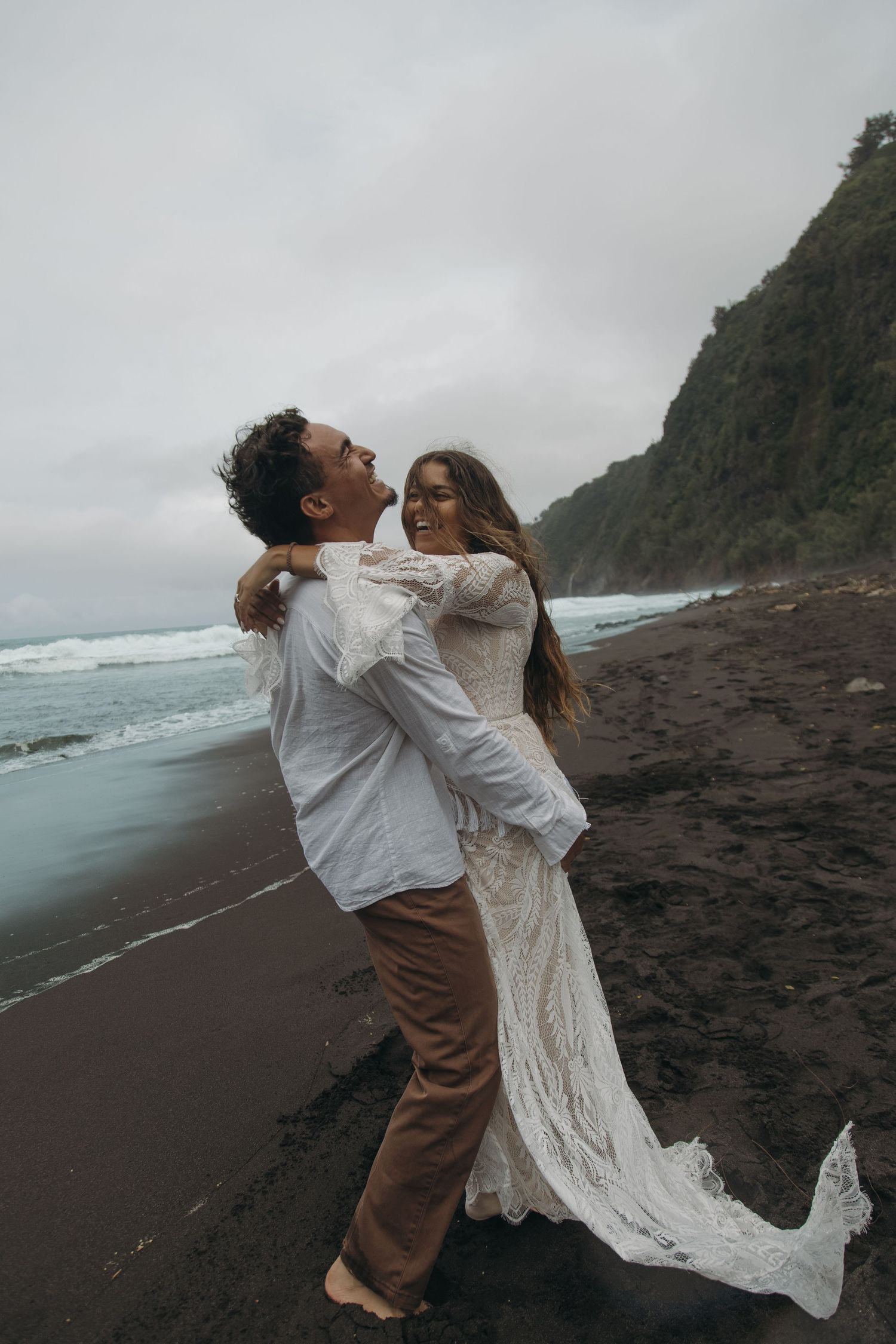 Couple embracing on black sand beach, bride in lace gown laughing as groom lifts her near ocean waves.