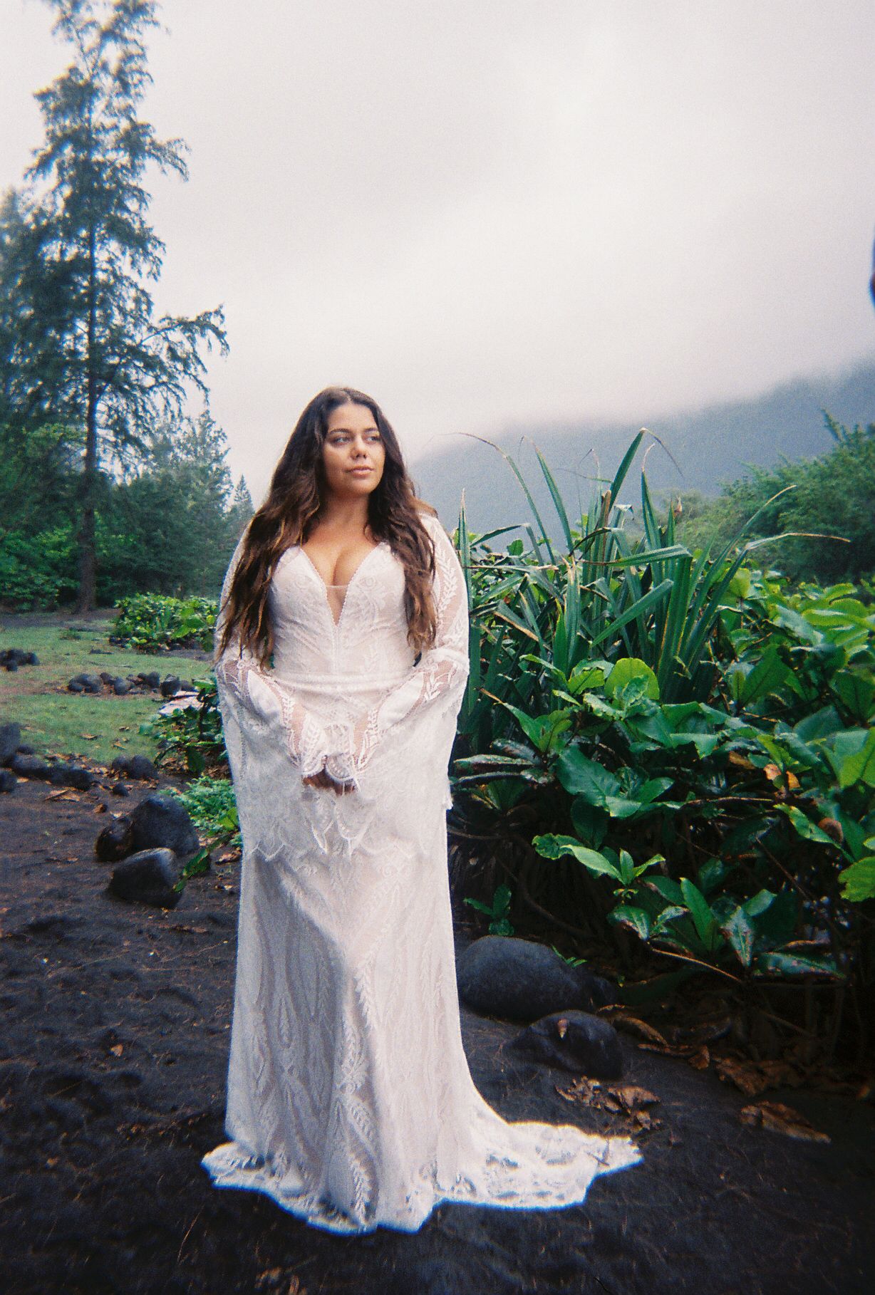 Woman in white lace boho wedding gown standing in lush tropical garden with misty mountains.
