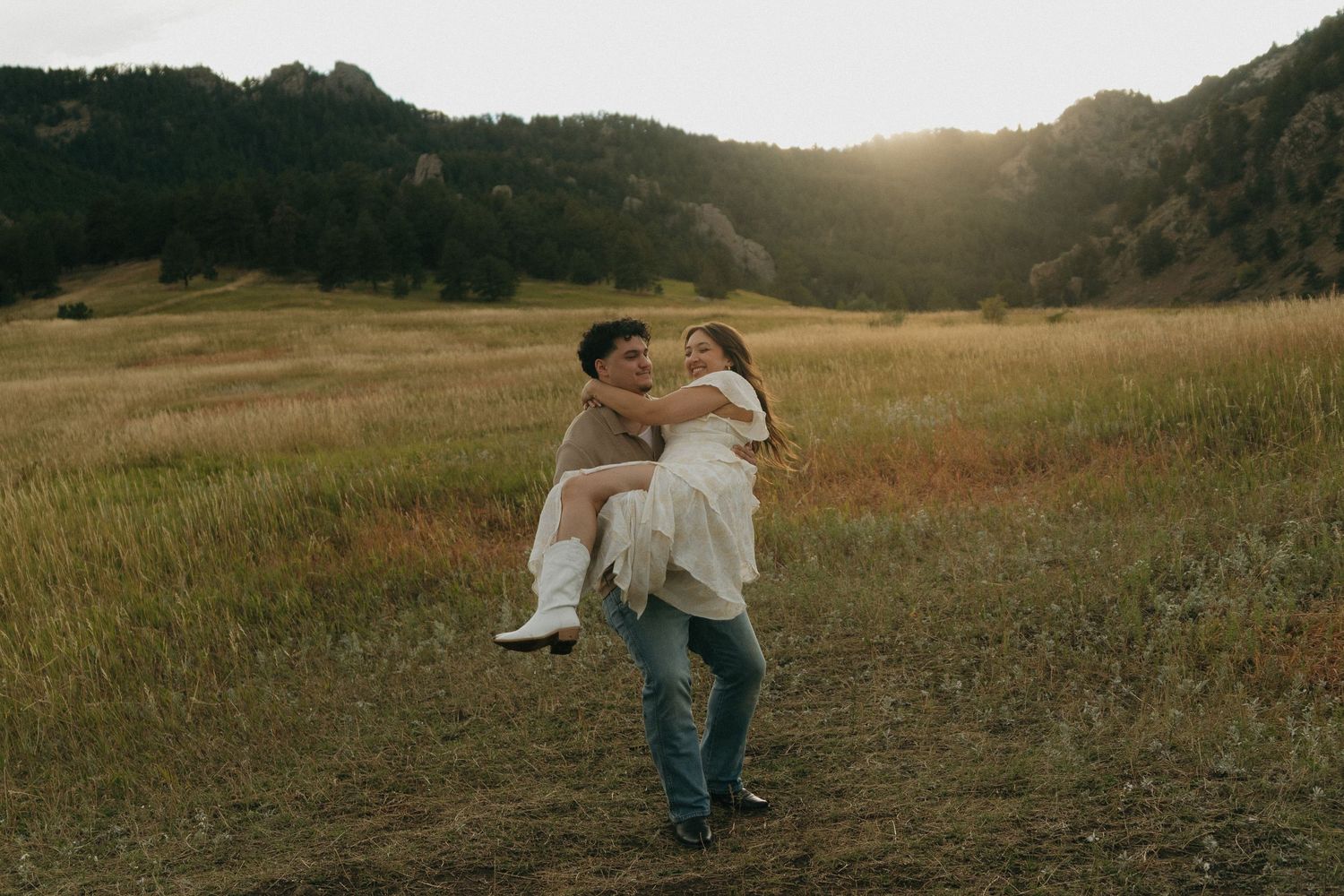 Man lifting woman in his arms in a sunlit mountain meadow during a romantic engagement photo session.