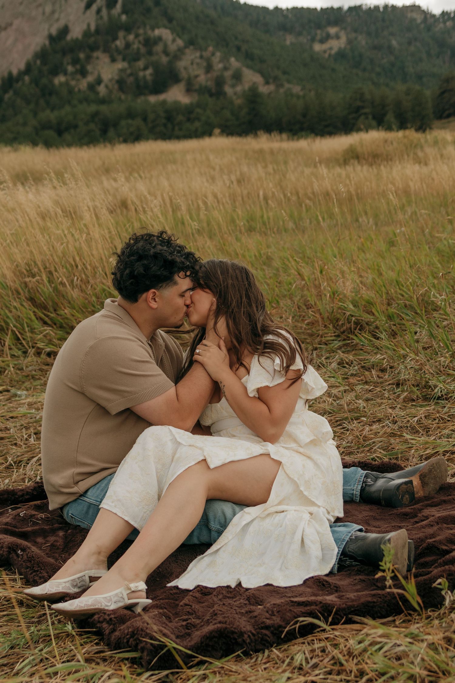 Romantic couple shares a kiss while sitting on a blanket in a scenic mountain meadow.