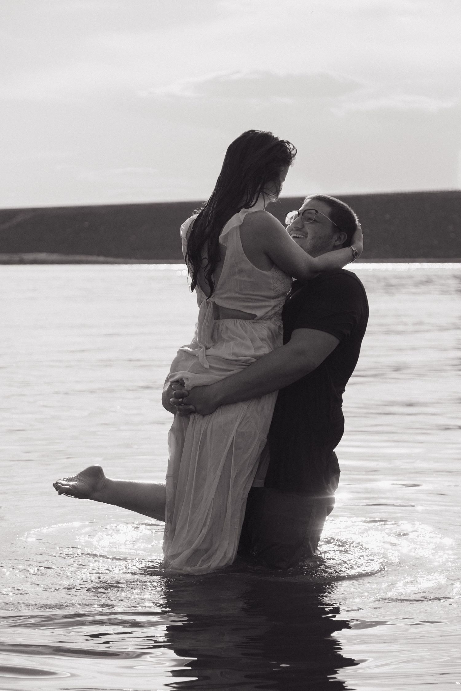Black and white photo of a couple embracing in water, man lifting woman in white dress near shoreline.