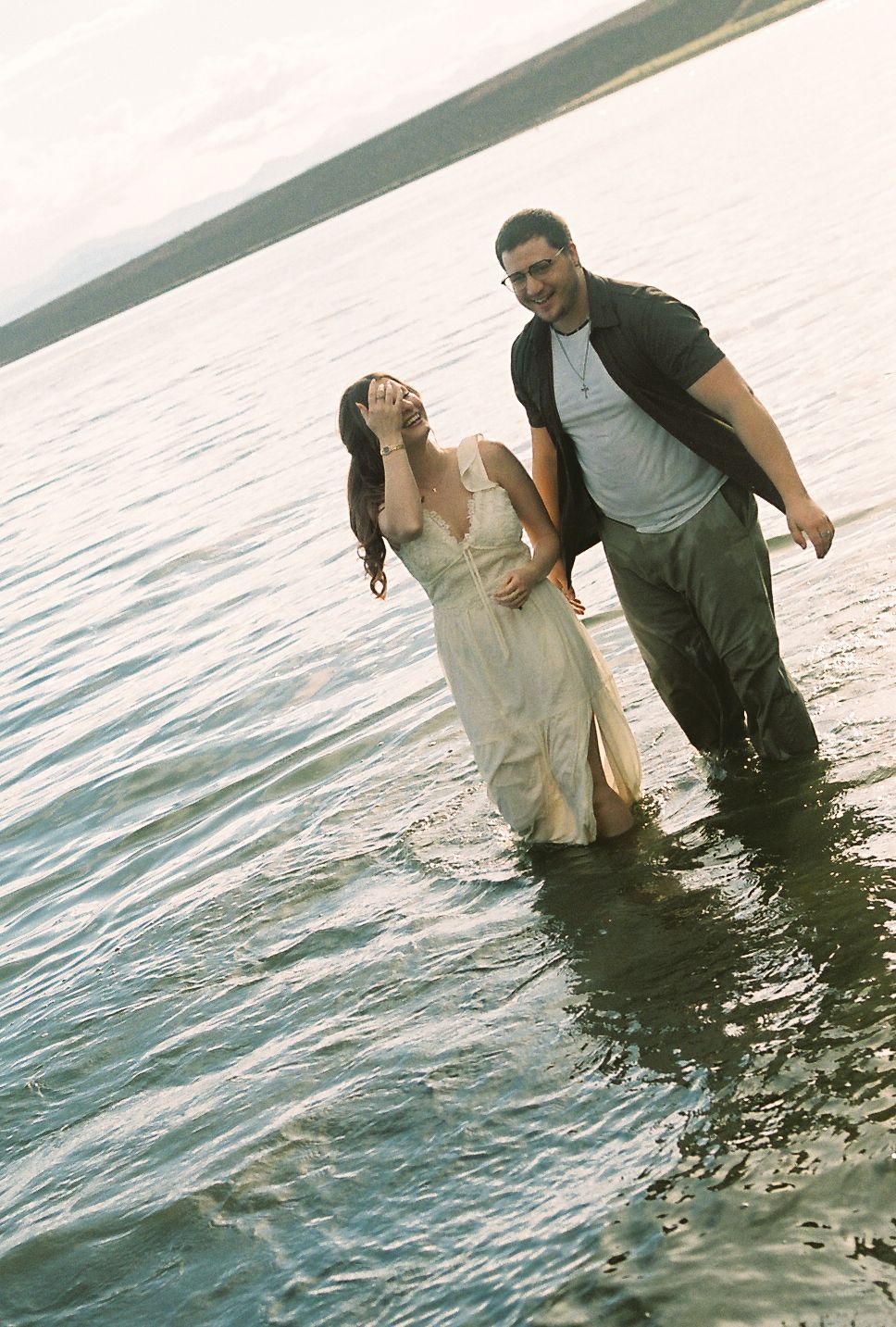 Couple laughing and walking hand in hand through shallow lake water, woman in white dress.