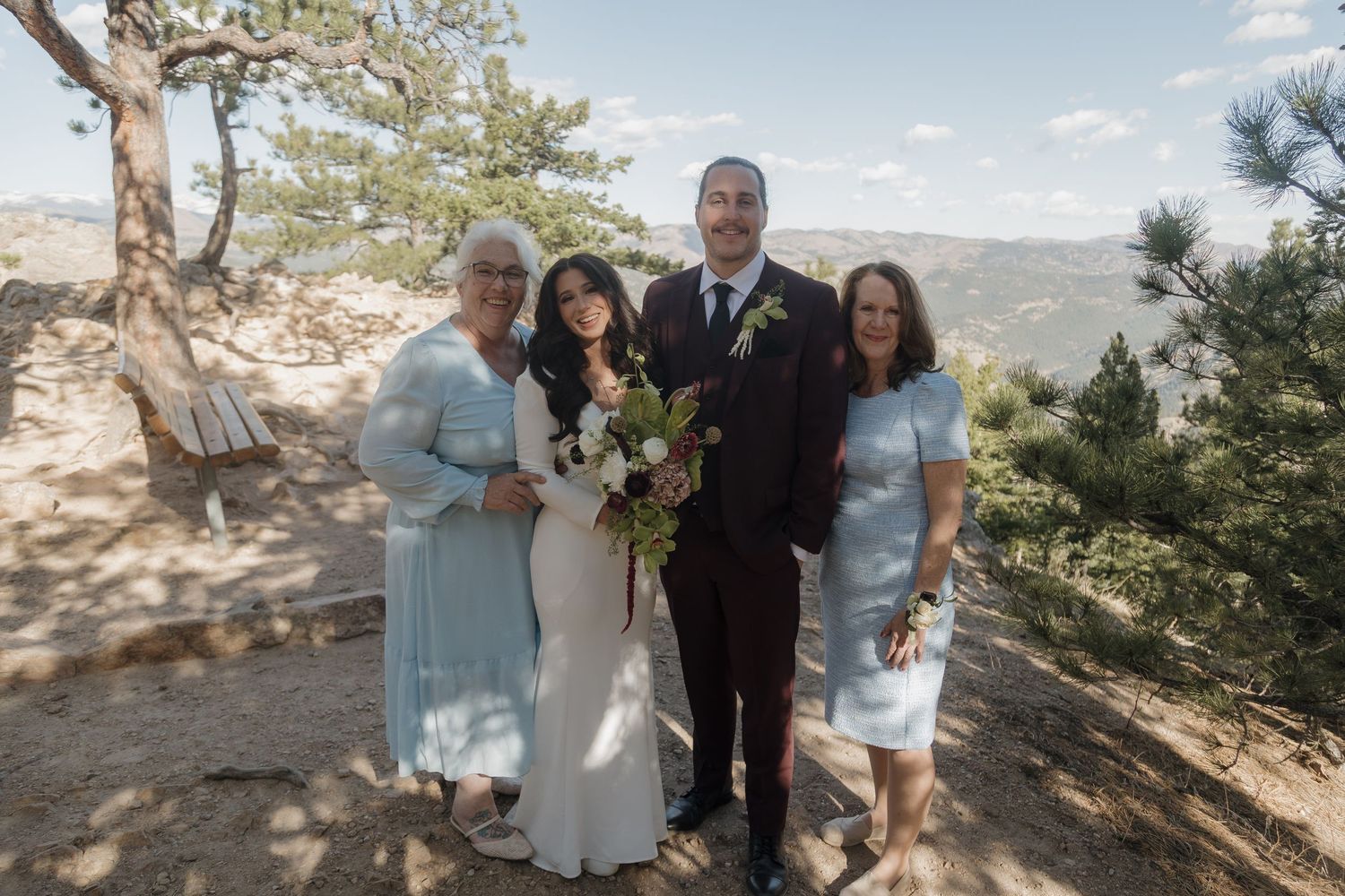 Bride and groom with two female guests in light blue dresses posing on mountain overlook with pine trees.