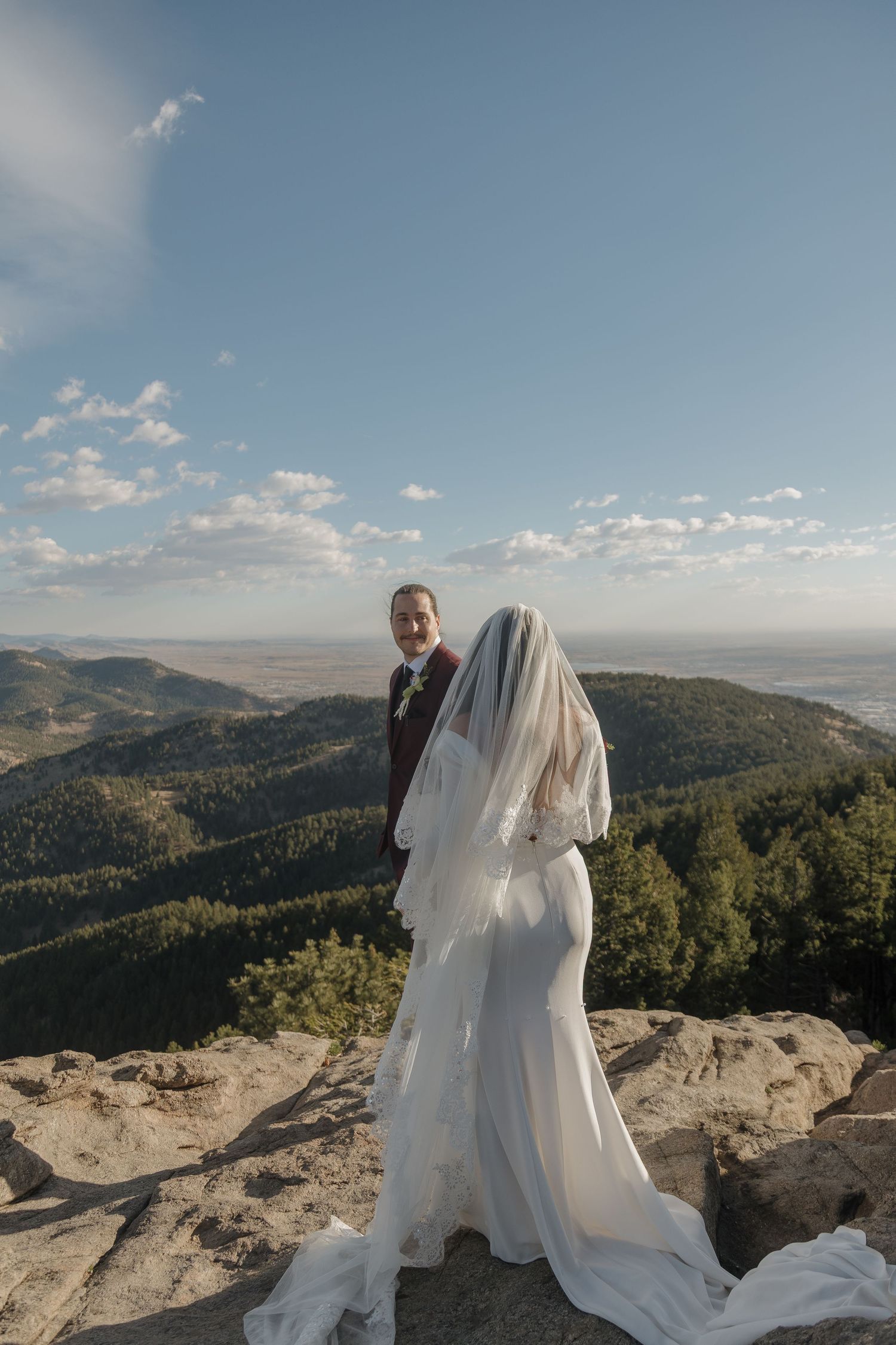 Bride in white gown with flowing veil and groom stand on mountain cliff with panoramic valley view.