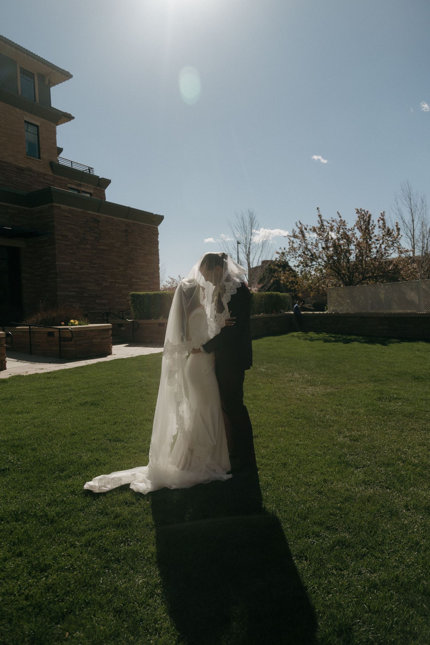 Bride and groom share a kiss on a sunny green lawn outside a brick wedding venue.