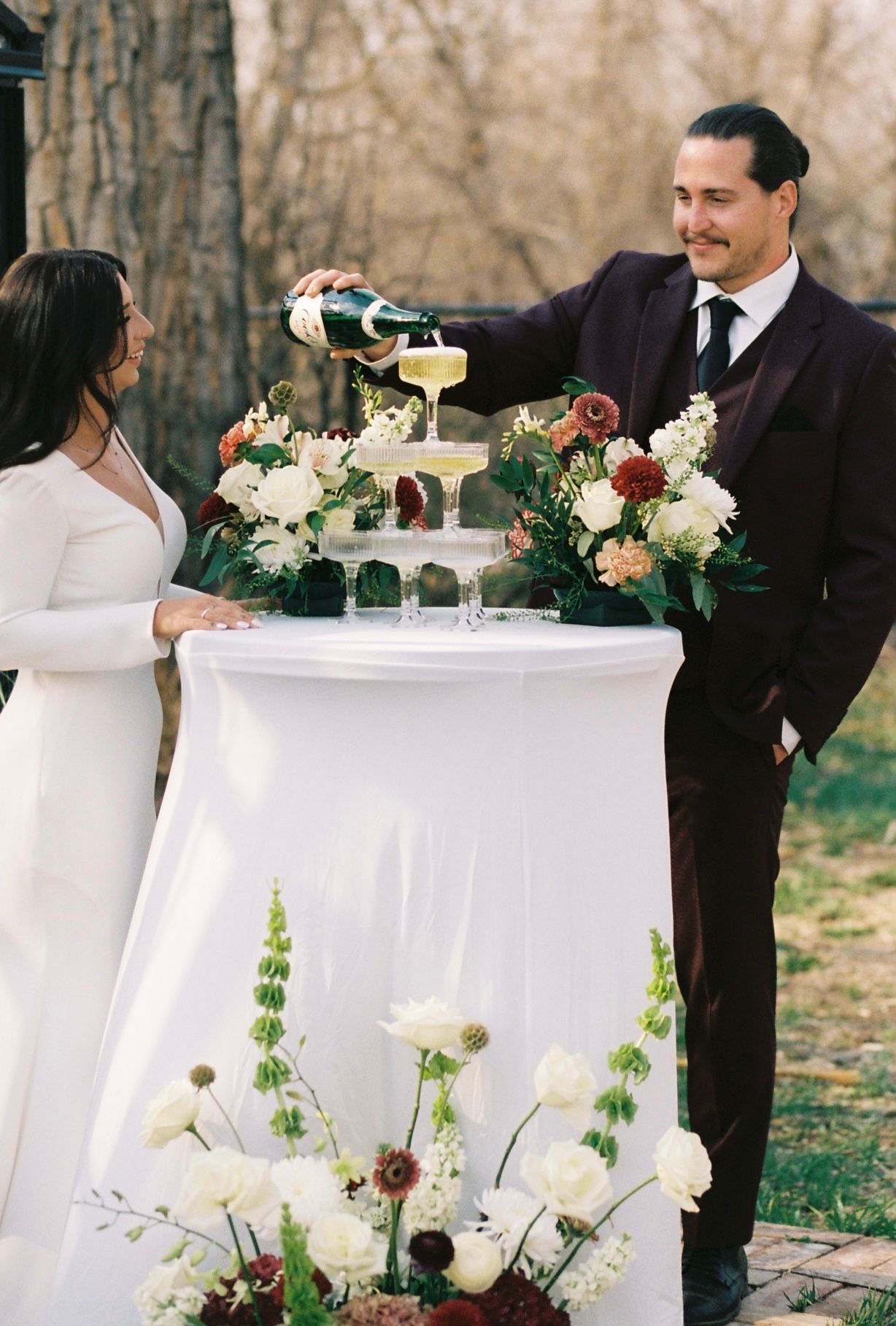 Groom in burgundy suit pouring champagne tower while bride watches at floral-decorated outdoor wedding reception.