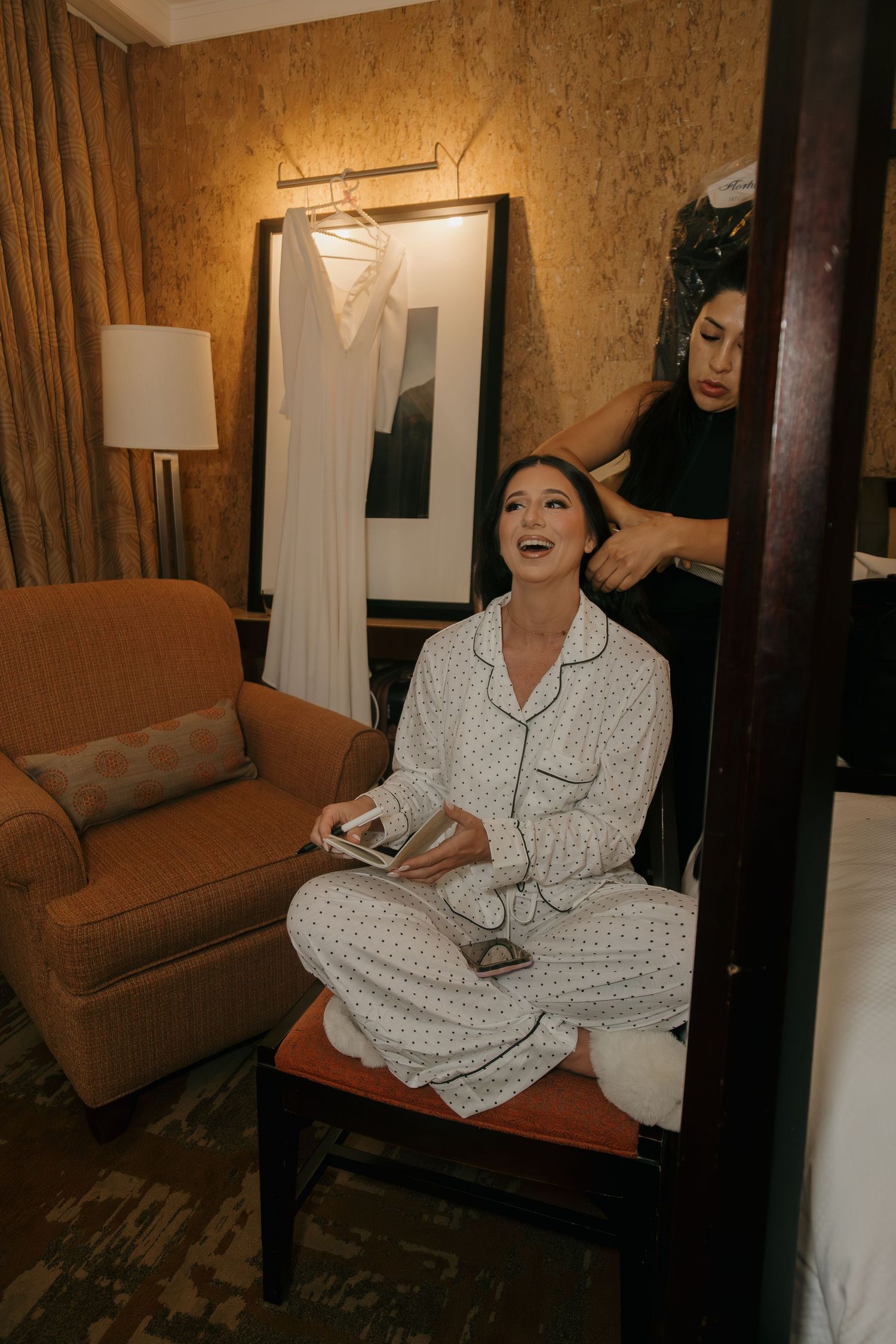 Bride in white pajamas laughing while getting hair done in a hotel room with wedding dress hanging in background.