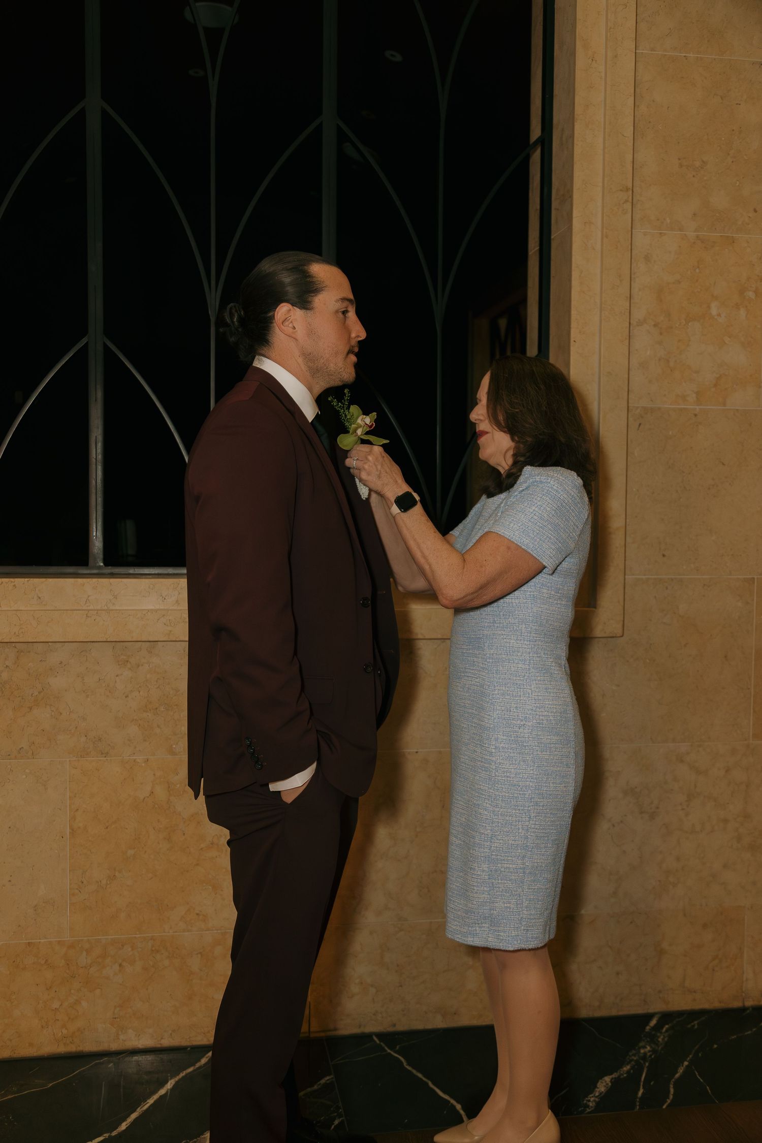 Woman in blue dress pins boutonniere on man in burgundy suit at formal event.