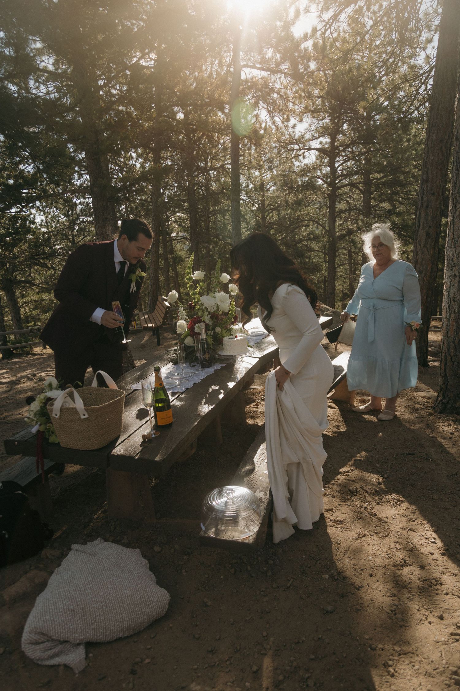 Bride and groom signing marriage certificate at sunlit outdoor picnic table reception in pine forest.