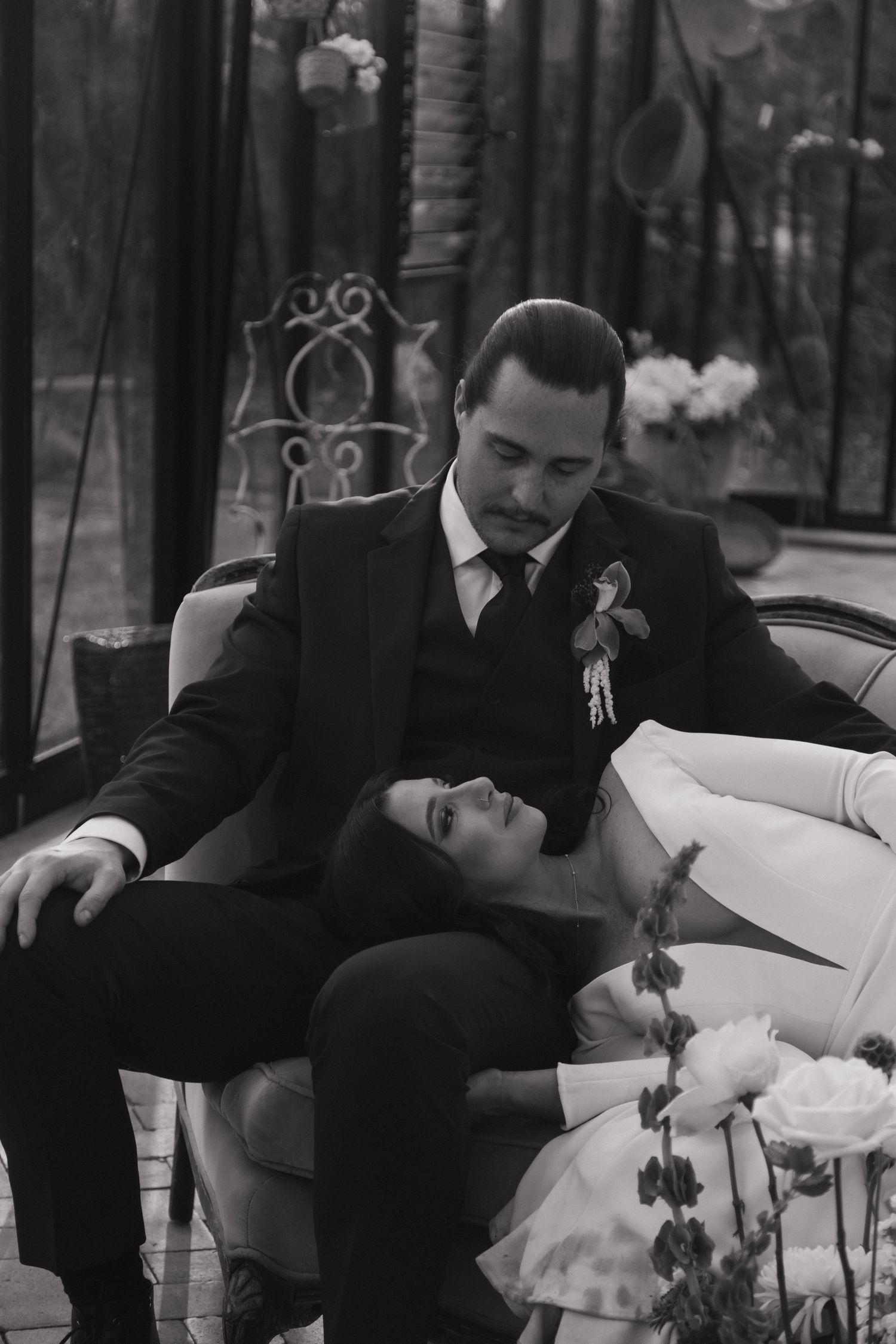 Groom in black suit holds bride in white dress on vintage sofa in romantic black and white portrait.