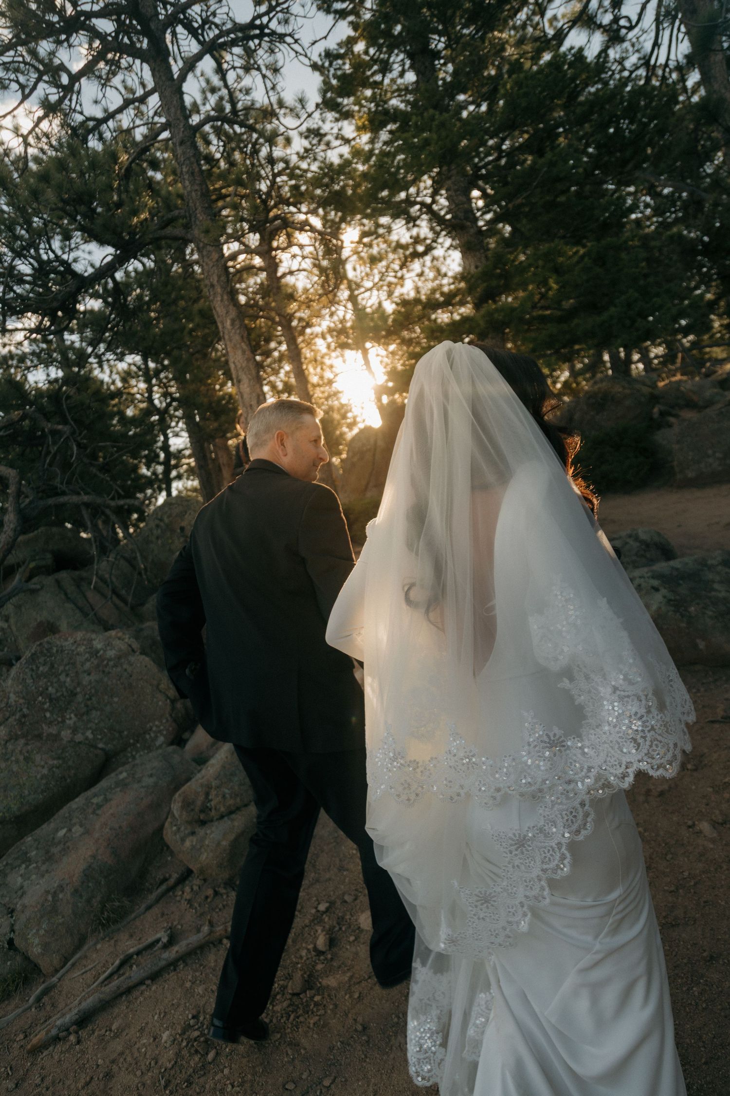 Bride and groom walk hand-in-hand through rocky pine forest toward golden sunset after ceremony.