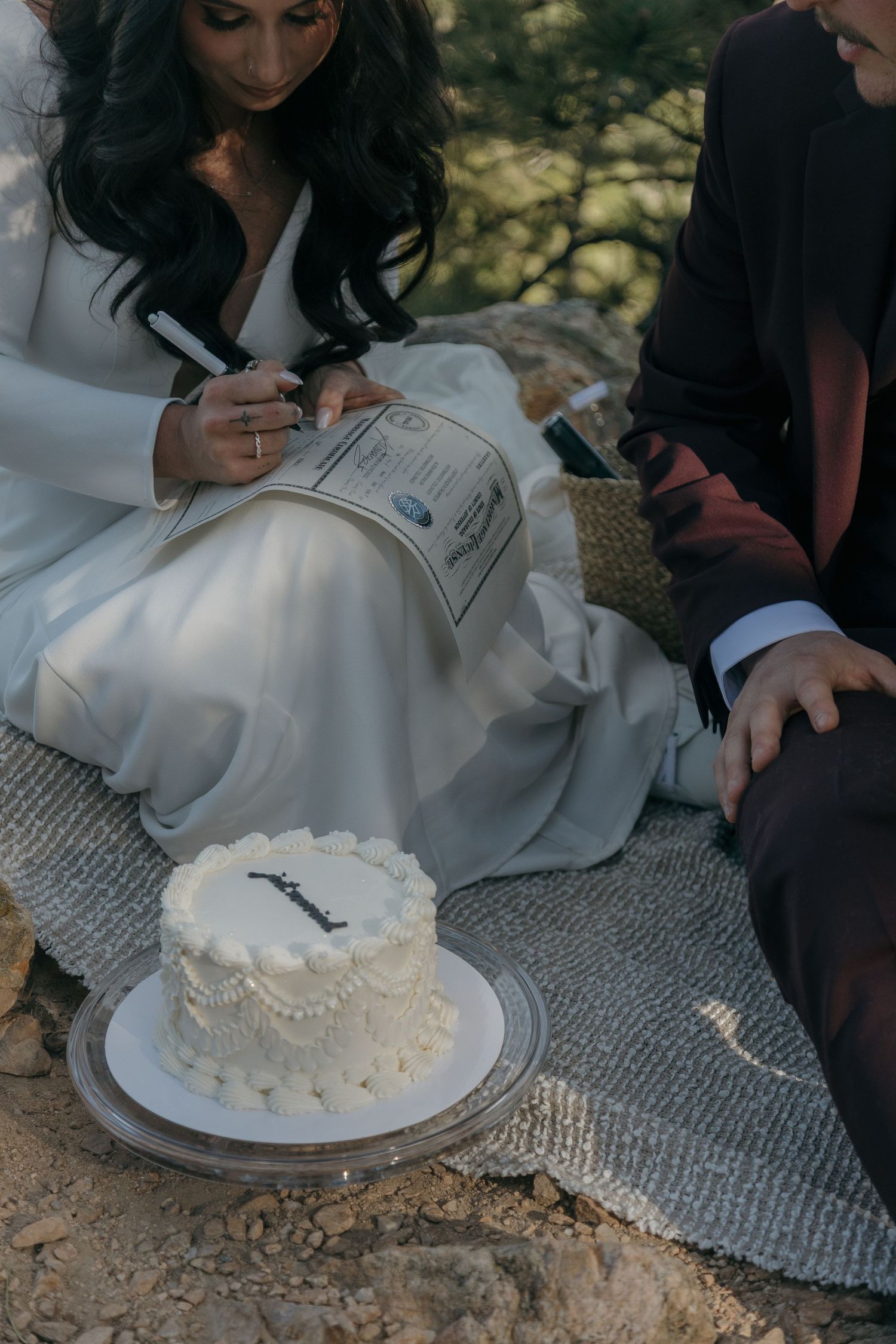 Bride and groom sign marriage certificate outdoors with small white wedding cake on blanket.