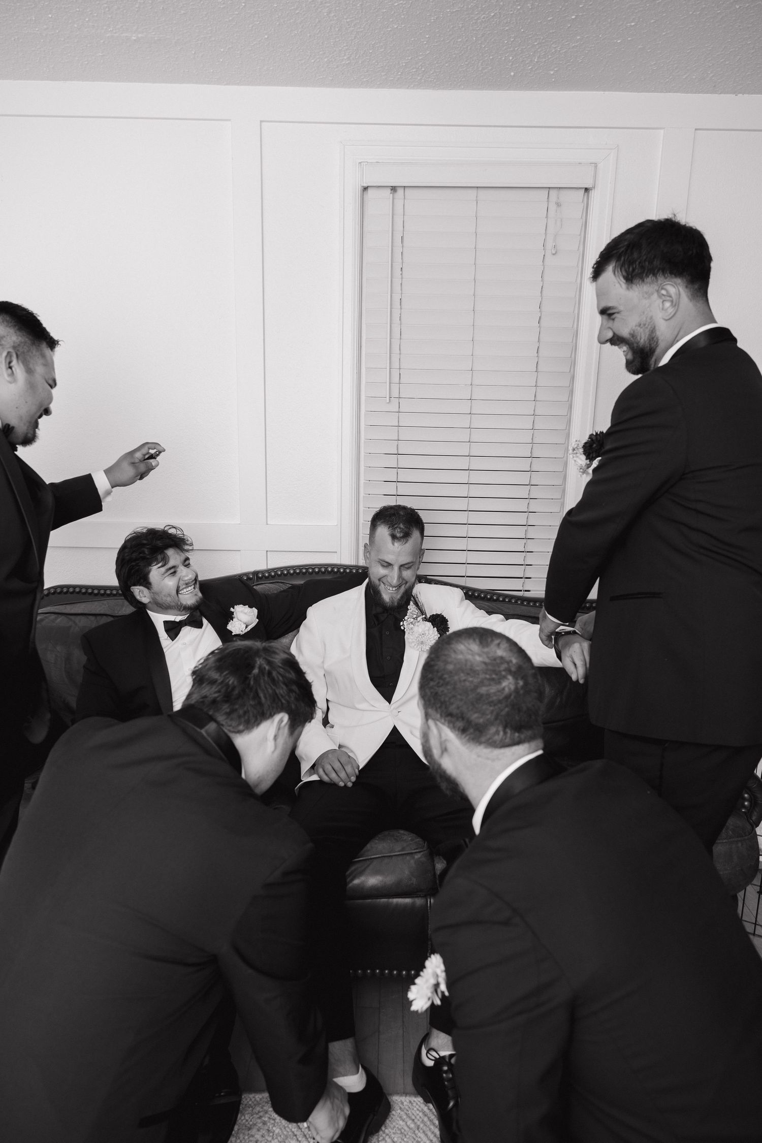 Groom in white tuxedo jacket surrounded by groomsmen in black suits during wedding preparations.