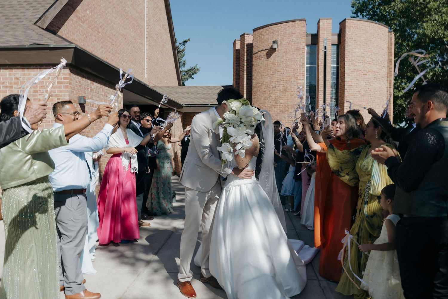 Newlywed couple kissing during joyful outdoor wedding ceremony exit with guests celebrating at brick church.