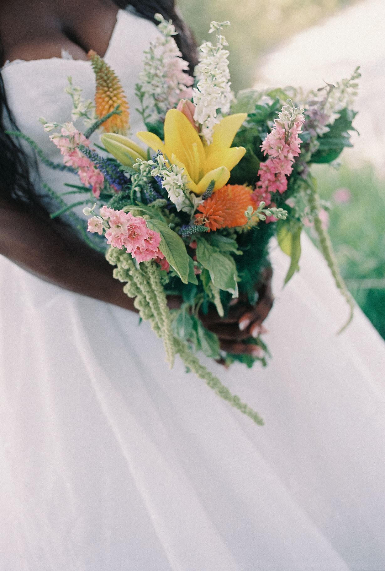 Bride in white gown holding vibrant wildflower bouquet with yellow lilies, pink blooms, and orange flowers.