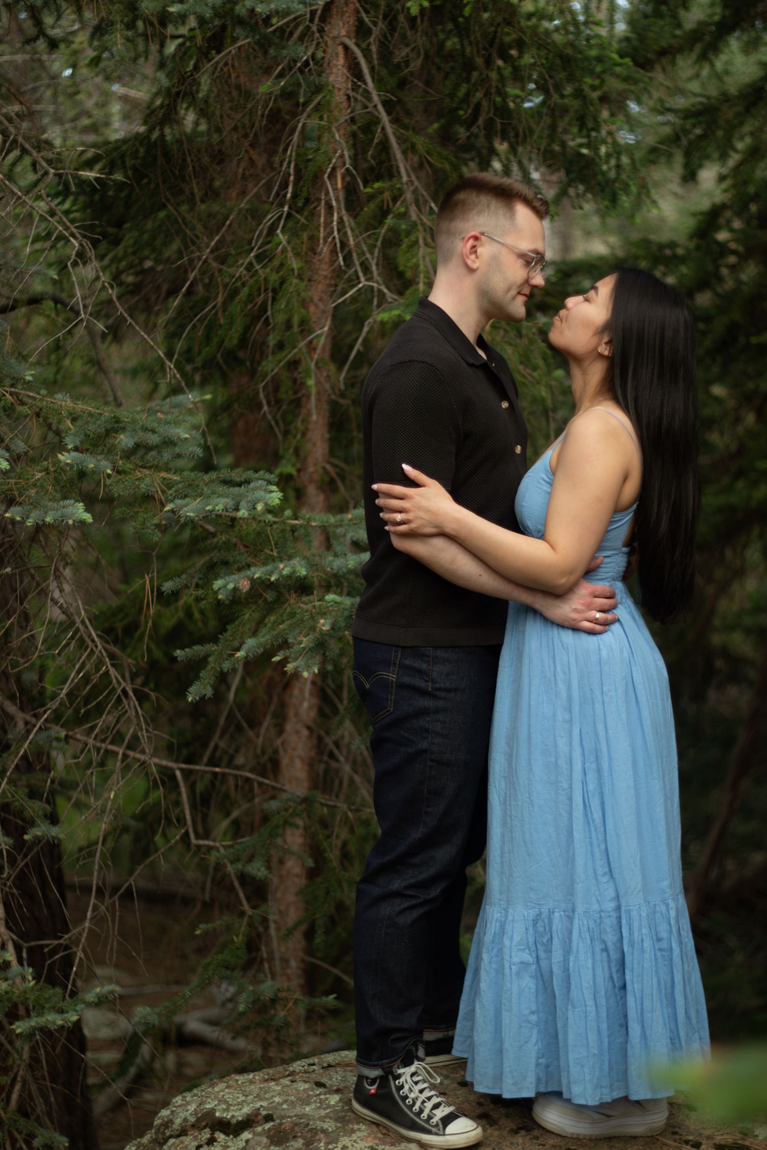 A couple in casual attire sharing a tender moment against a backdrop of pine trees.