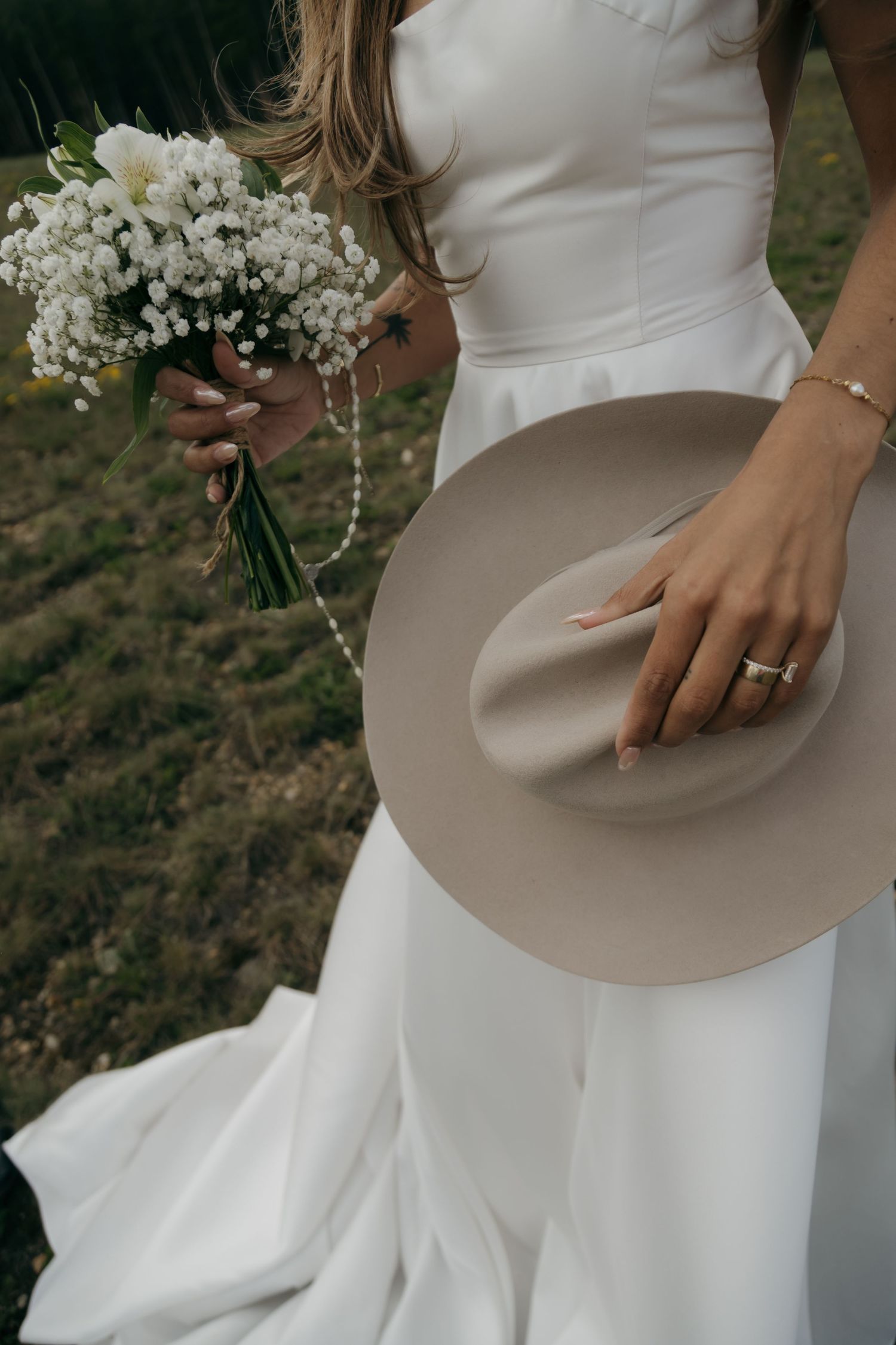 Bride holding white floral bouquet and beige wide-brim hat wearing wedding ring in white gown.
