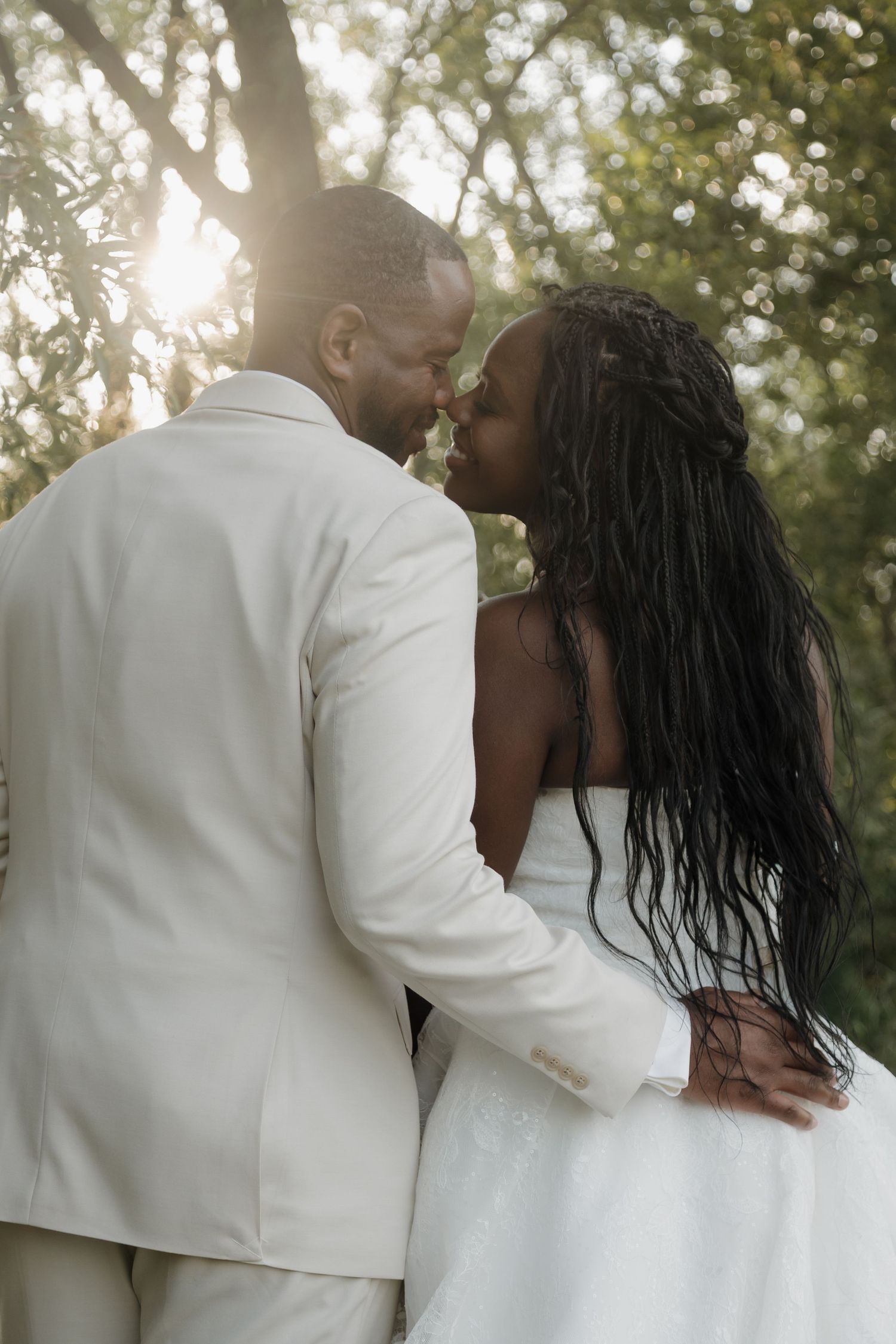Black couple in wedding attire sharing a tender kiss outdoors with sunlight filtering through trees.