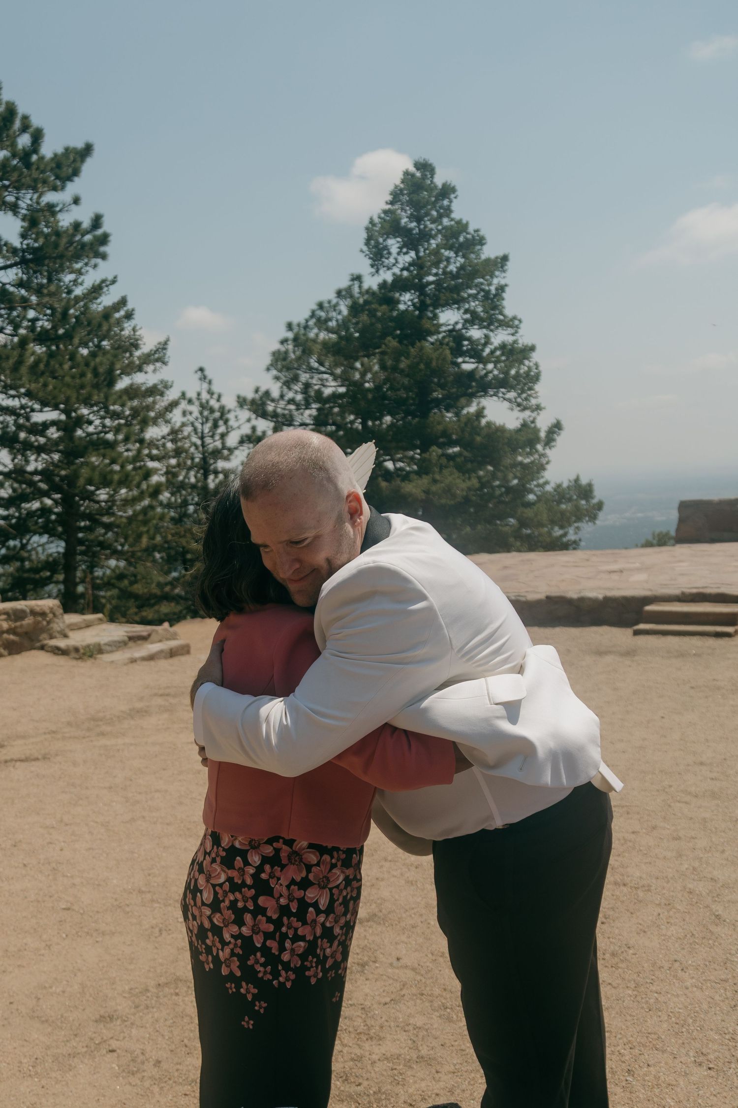 Groom in white jacket sharing an emotional hug with a guest at an outdoor wedding.