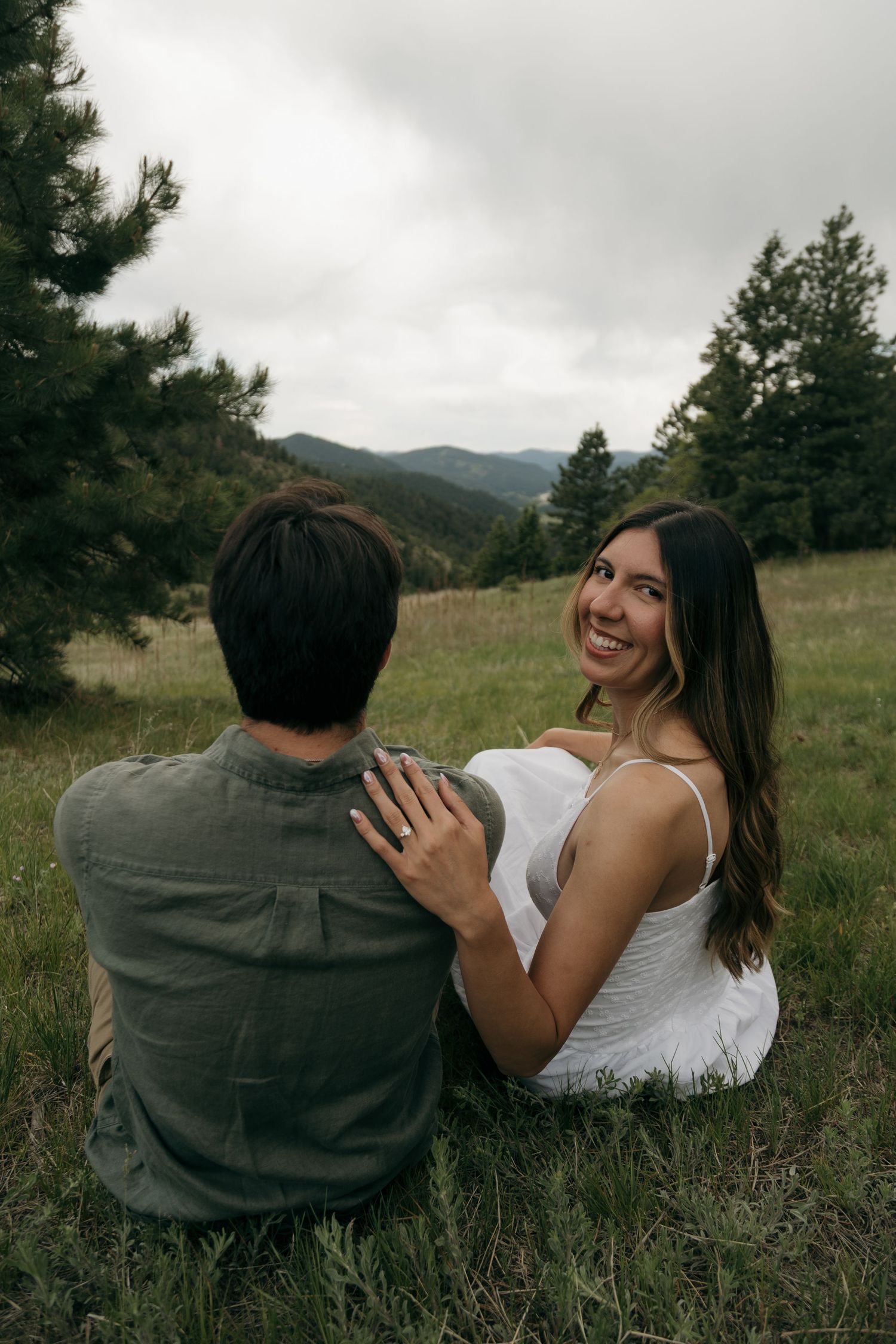 A smiling woman in a white dress sits with her partner in a lush mountain meadow surrounded by evergreen trees.