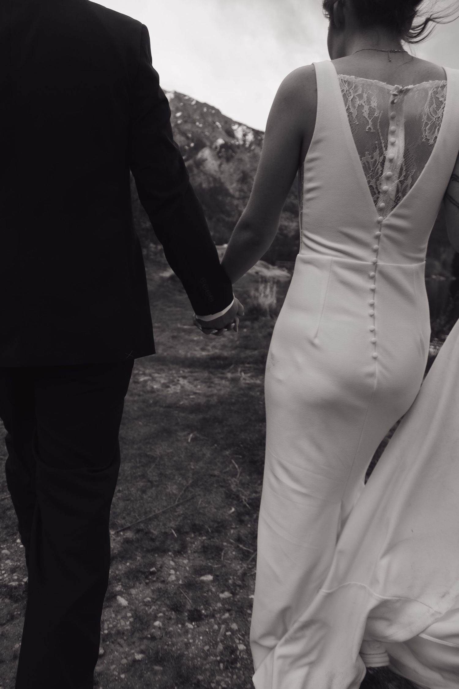 Black and white close-up of bride and groom holding hands, showcasing lace back detail of wedding dress.
