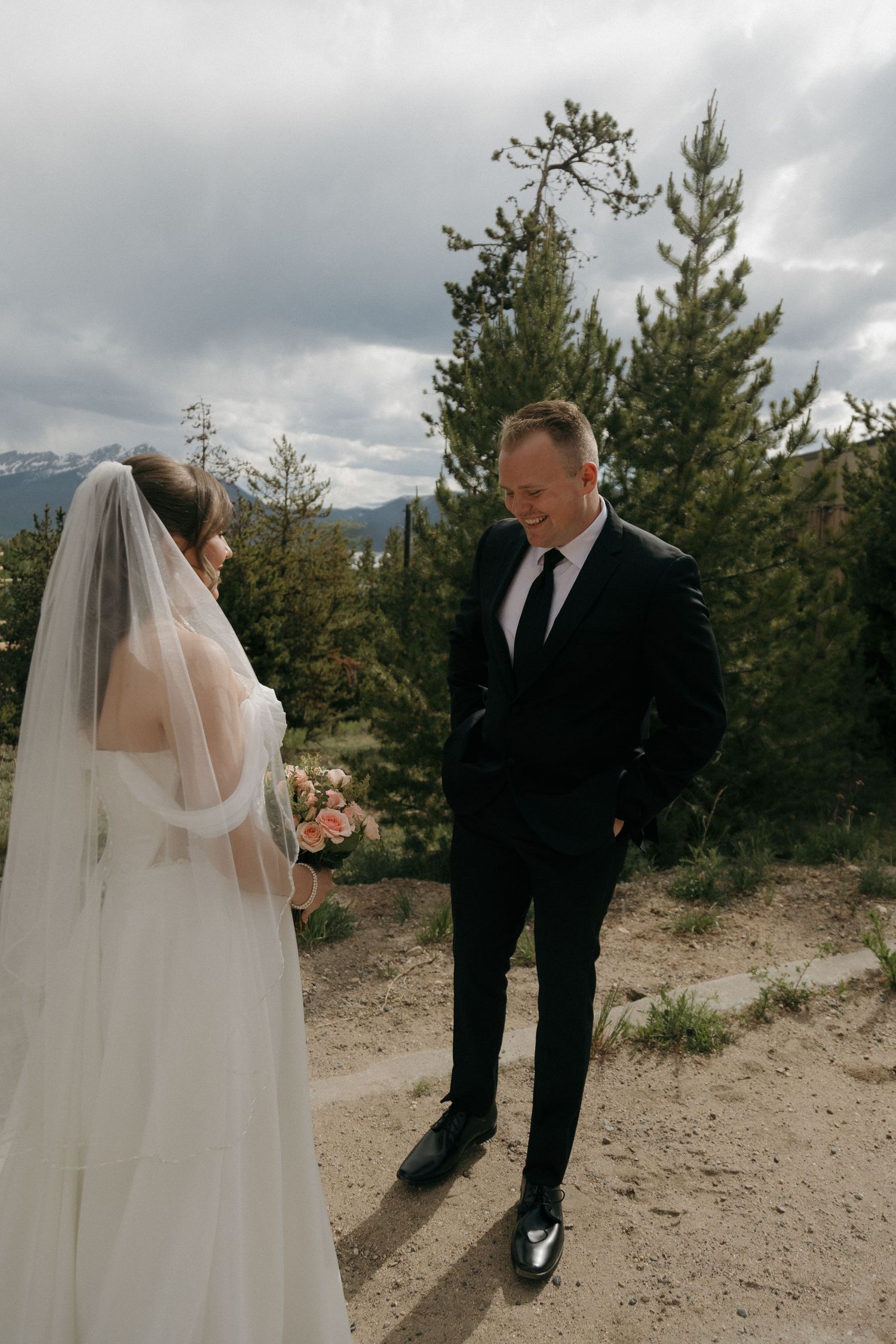 Groom in black suit smiles at veiled bride holding pink bouquet on a mountain trail during first look.