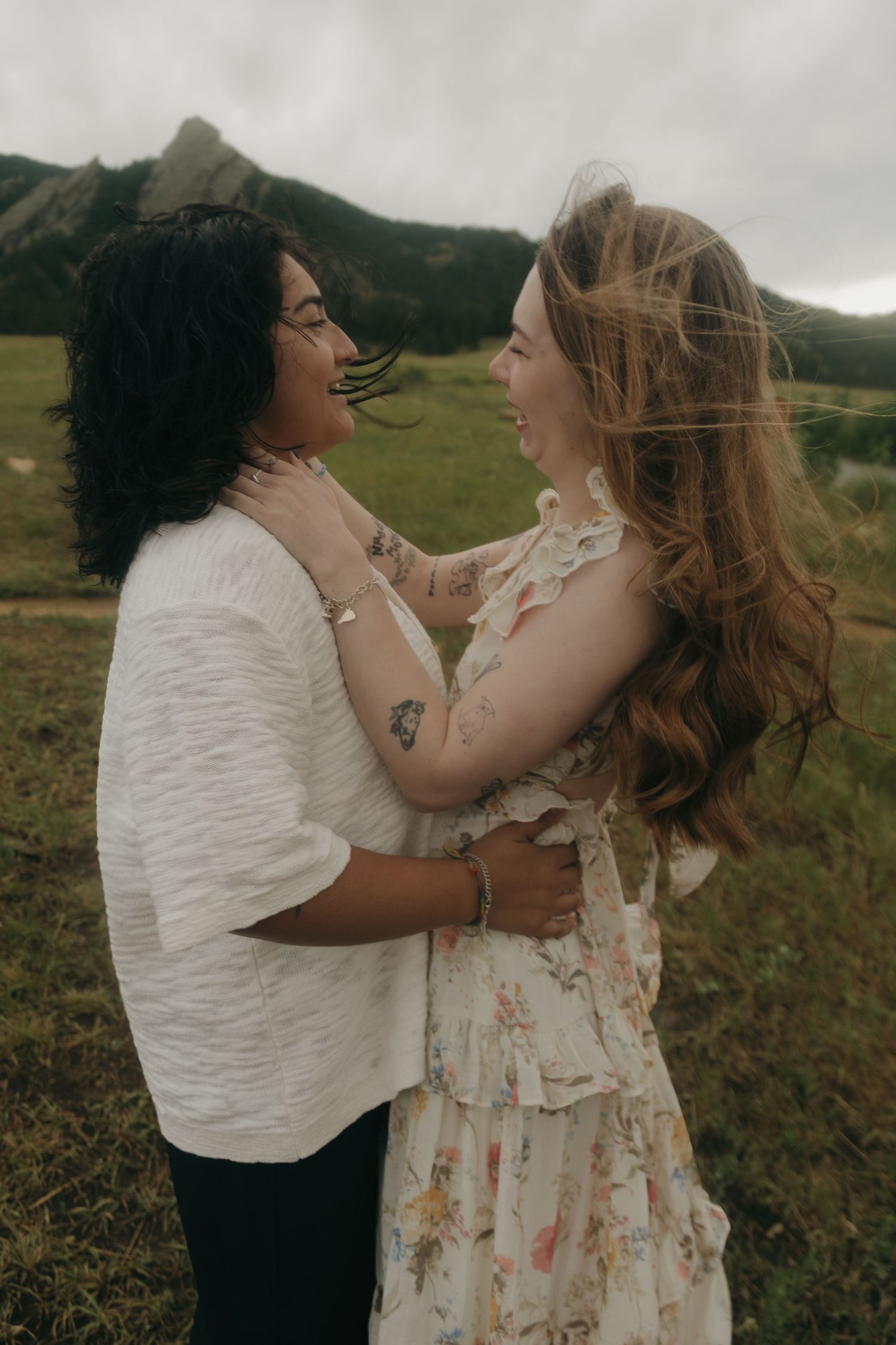 Two women embrace tenderly in a green mountain meadow, smiling at each other on a windy day.