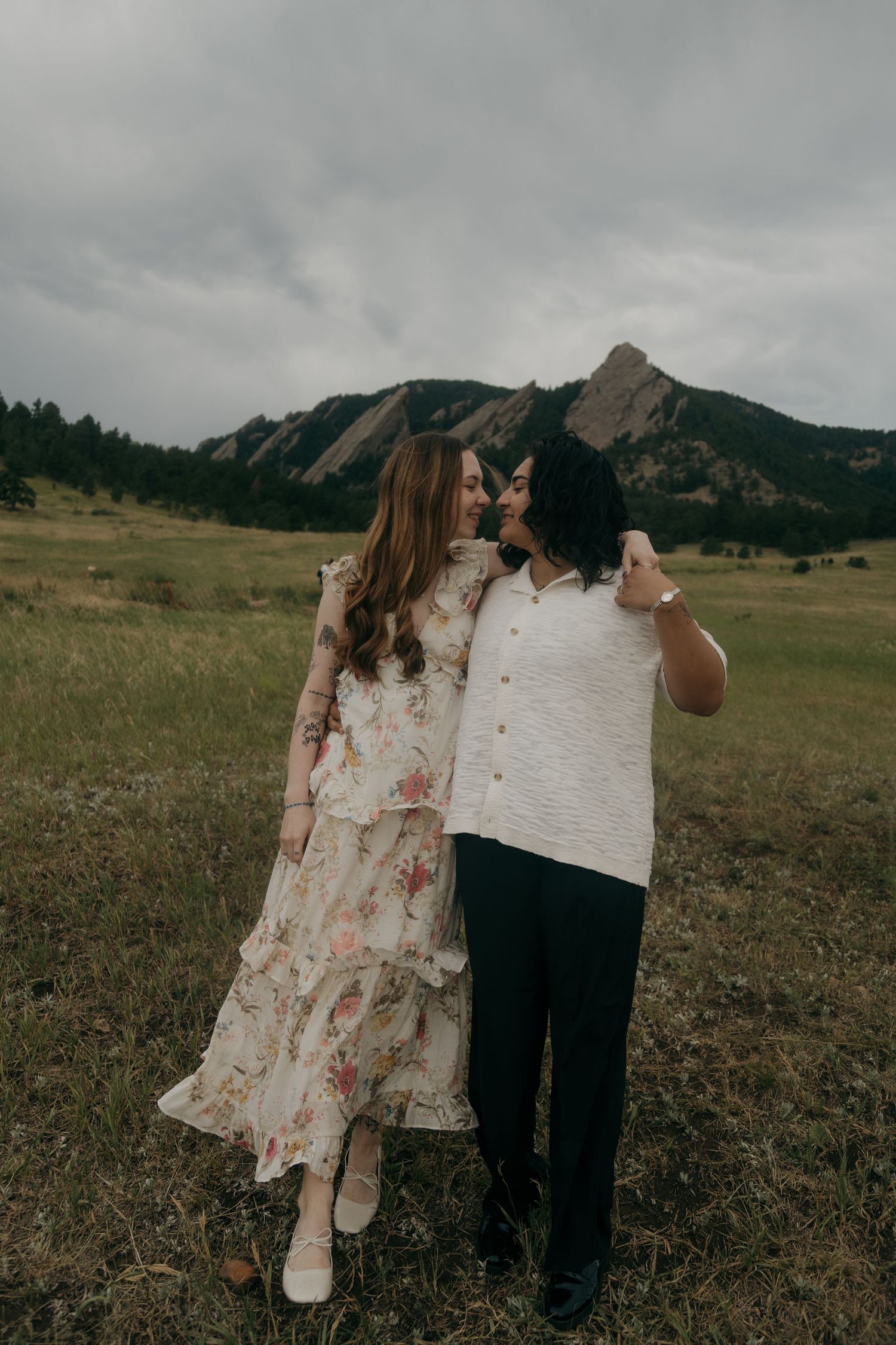 Couple embracing in a meadow with the Flatirons rock formations in background under cloudy skies.
