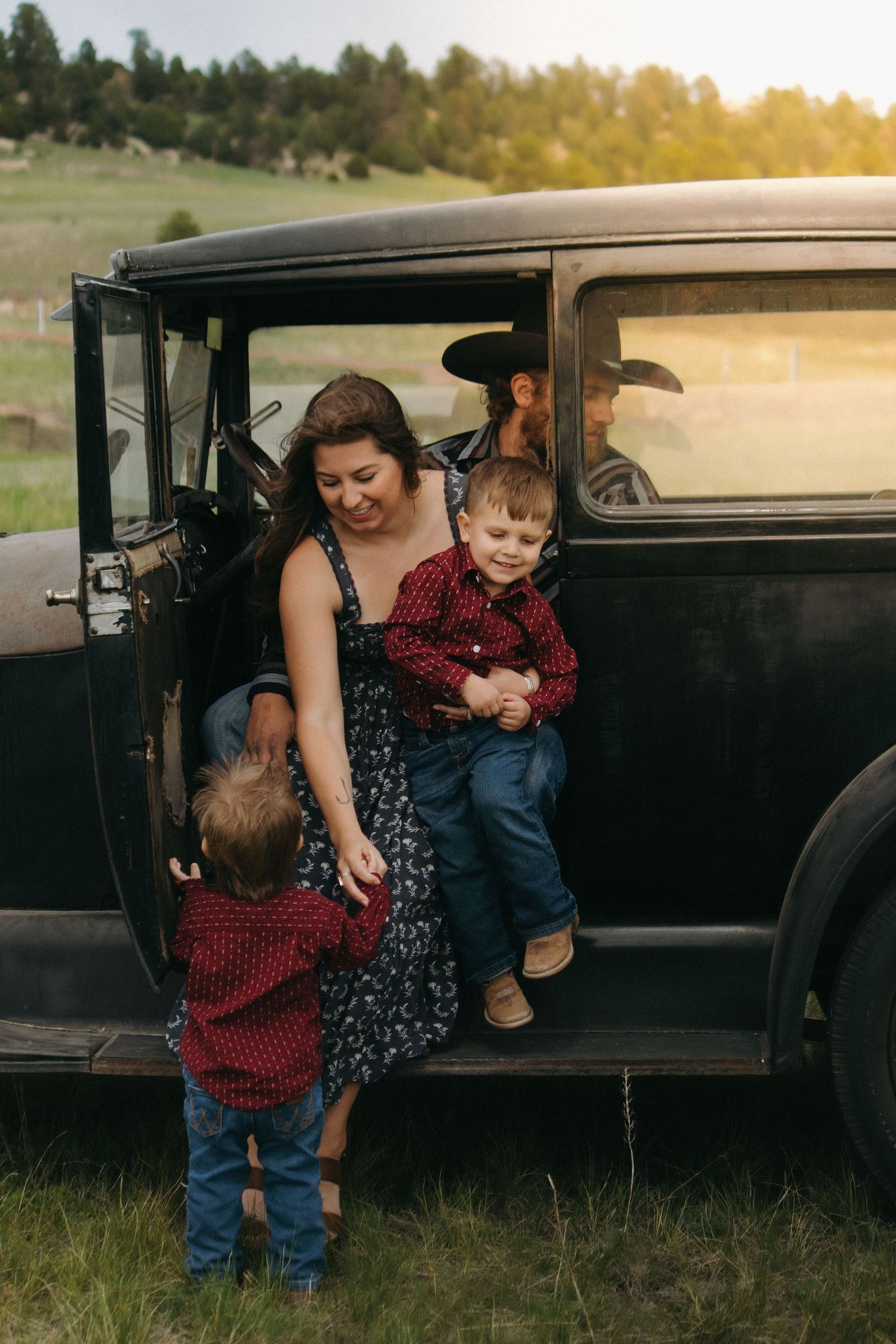 Family gathering around a classic black car in a scenic field during golden hour.